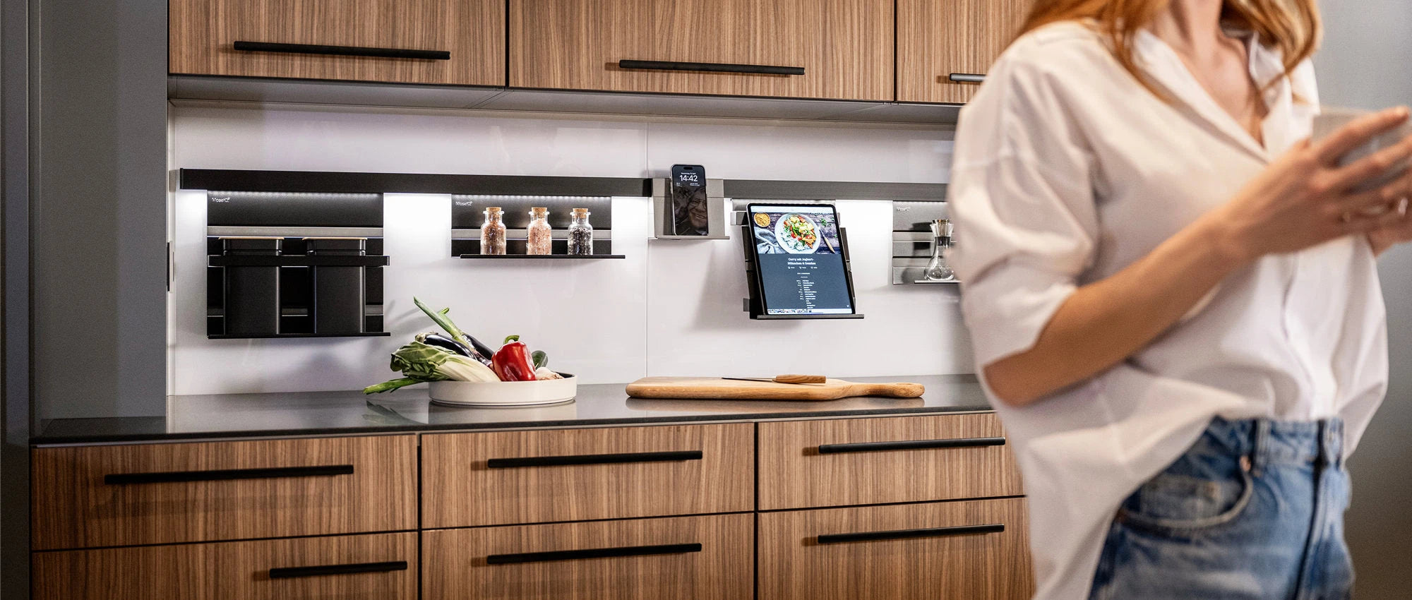 Eine moderne Küche mit Holzschränken, einem Schneidebrett, Gemüse auf einem Tablett, Gewürzen und einem Tablet, das an der Spüle befestigt ist. Eine Person in weißem Hemd und Jeans steht im Vordergrund, leicht unscharf.