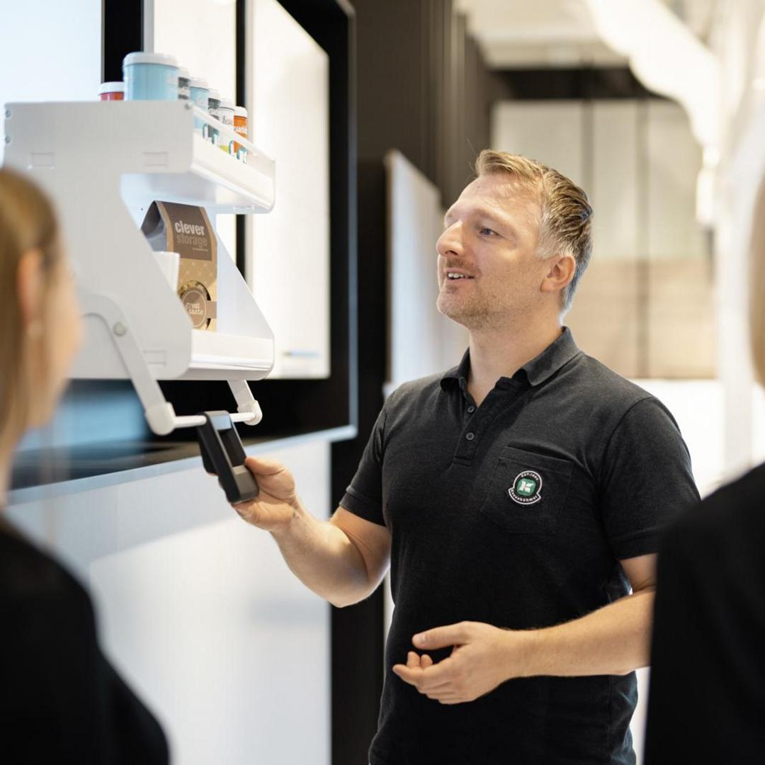 Ein Mann in einem schwarzen Polohemd demonstriert zwei Personen in einem modernen, gut beleuchteten Raum einen an der Wand montierten Automaten. Er lächelt und hält eine schwarze Karte in die Nähe des Automaten.