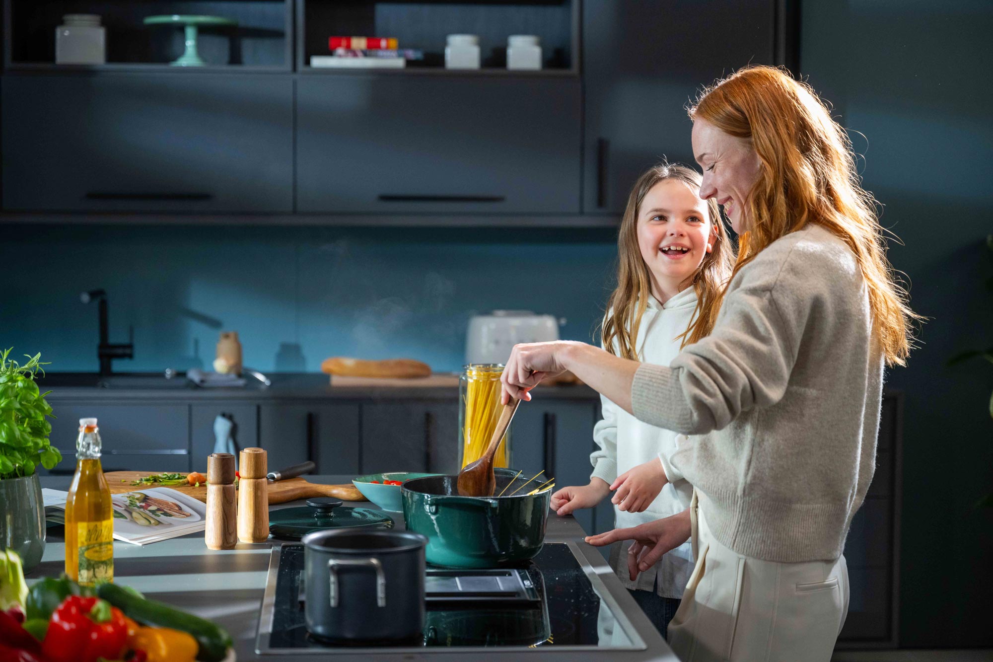 Eine Frau und ein junges Mädchen lächeln und kochen gemeinsam in einer modernen Küche. Sie geben Spaghetti in einen Topf auf dem Herd, auf dem Tresen stehen frisches Gemüse und Olivenöl.