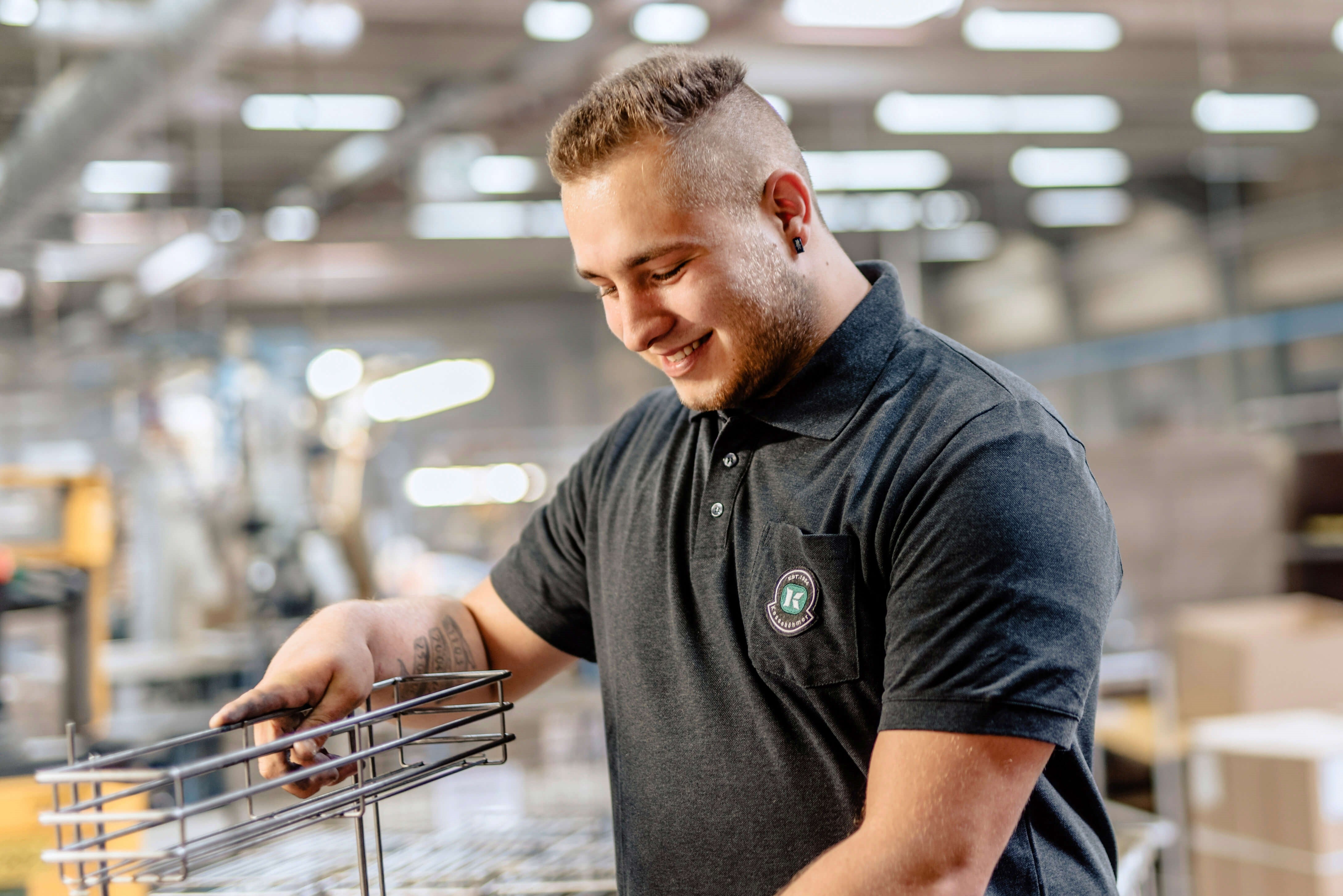 Eine lächelnde Person in einem dunklen Polohemd arbeitet an einem Metallgestell in einer hell erleuchteten Industrie- oder Produktionsumgebung.