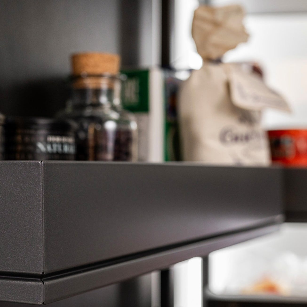 Nahaufnahme eines Kühlschranktürregals mit verschiedenen Gegenständen, darunter ein Glas mit Korkdeckel, eine Papiertüte und Konserven, wobei das Innere des Kühlschranks im Hintergrund zu sehen ist.