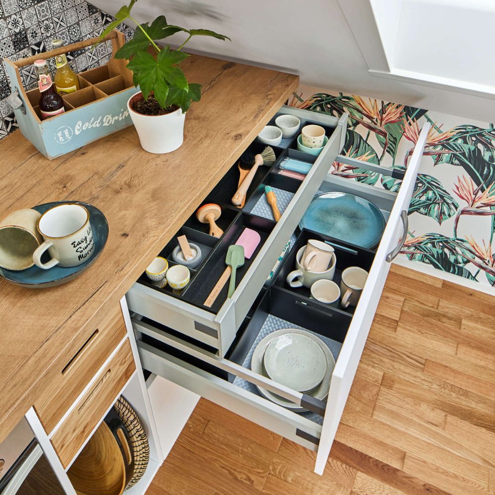 In den geöffneten Küchenschubladen sind Utensilien, Teller, Schüsseln und Tassen übersichtlich angeordnet. Eine Pflanze und ein Tablett mit Gewürzen stehen auf der hölzernen Arbeitsplatte. Der Boden und die Wände sind mit bunten Mustern versehen.