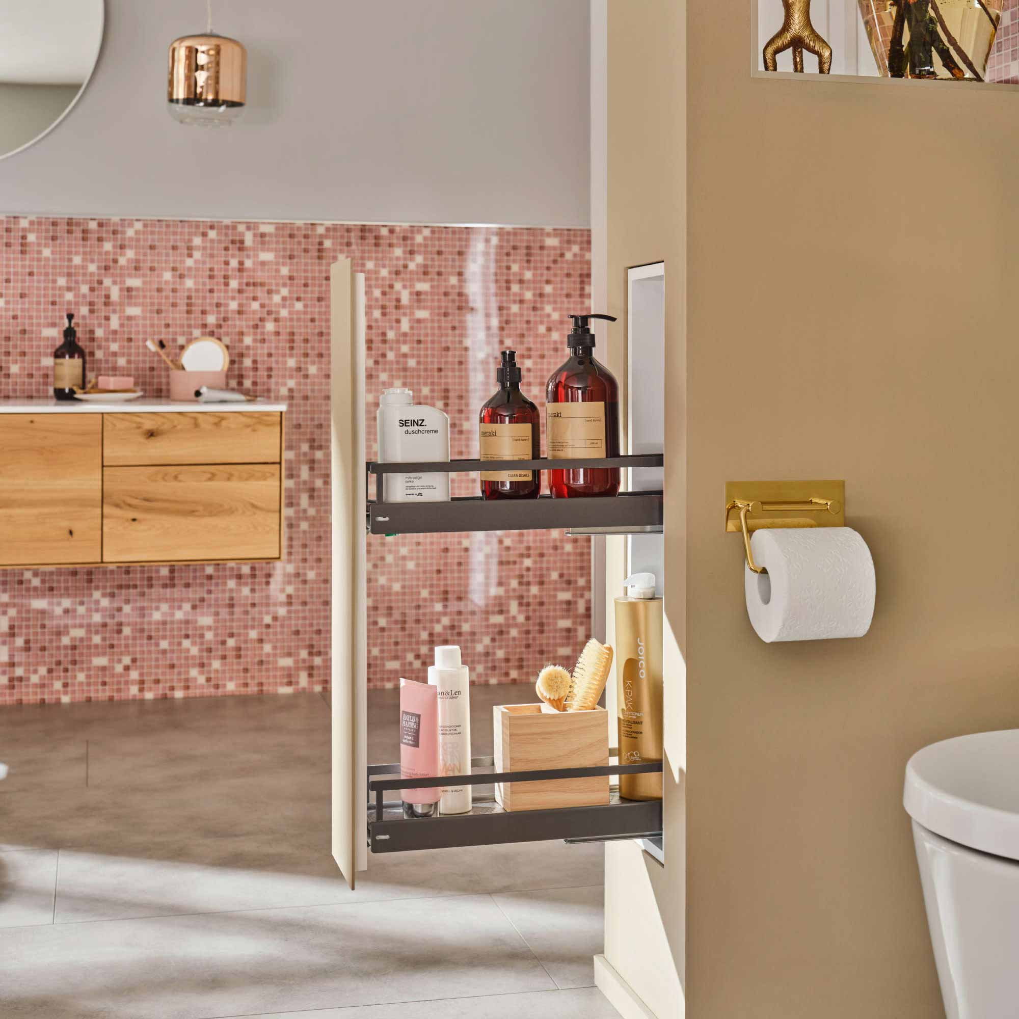 Ein modernes Badezimmer mit rosafarbenen Mosaikfliesen, einem hölzernen Waschtisch und einem ausziehbaren Regal mit Toilettenartikeln. Ein Toilettenpapierhalter ist an einer beigen Wand neben einer weißen Toilette angebracht.