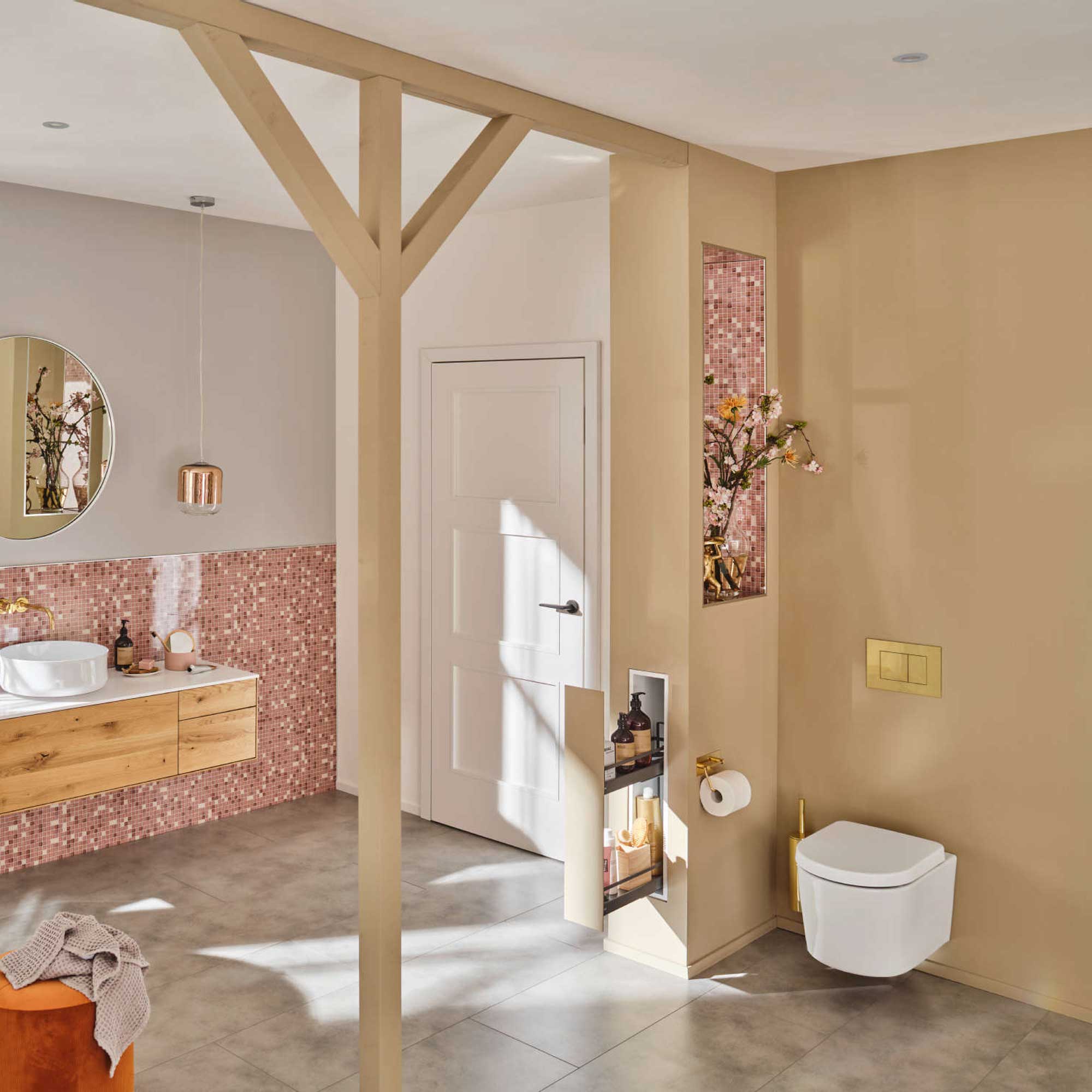 Modernes Badezimmer mit rosafarbenen Mosaikfliesen, schwebendem Holzwaschtisch, rundem Spiegel, wandmontierter Toilette und Einbauregalen mit Dekoration. Neutrale Wände und Böden mit natürlichem Lichteinfall.