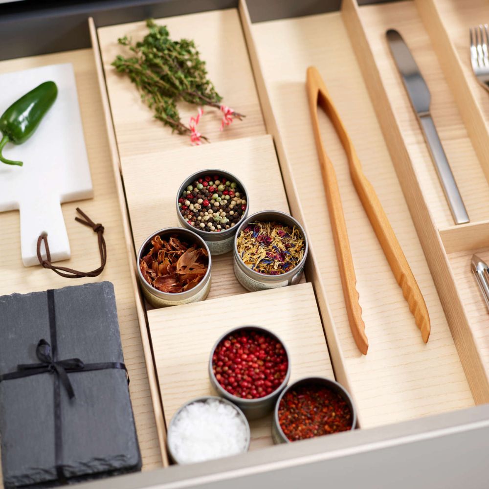 In einer organisierten Küchenschublade steht die FineLine LiniQ Gewürzeinlage - Gewürz Organizer, gefüllt mit Gewürzdosen, Holzzangen, Besteck, frischen Kräuterbündeln, einem grünen Chili auf einer weißen Tafel und schwarzen Schieferuntersetzern, die mit einer Schnur zusammengebunden sind.
