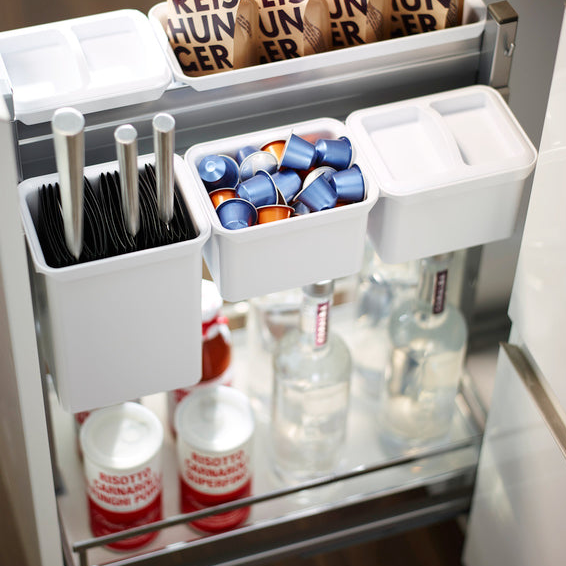 Die YouboXx Einhängeboxen - Set 1 für DISPENSA ordnen in der Schublade Messer, Einwegbesteck und Kaffeepads. Darunter befinden sich Flaschen mit klarer Flüssigkeit und Dosen mit roten und weißen Etiketten.