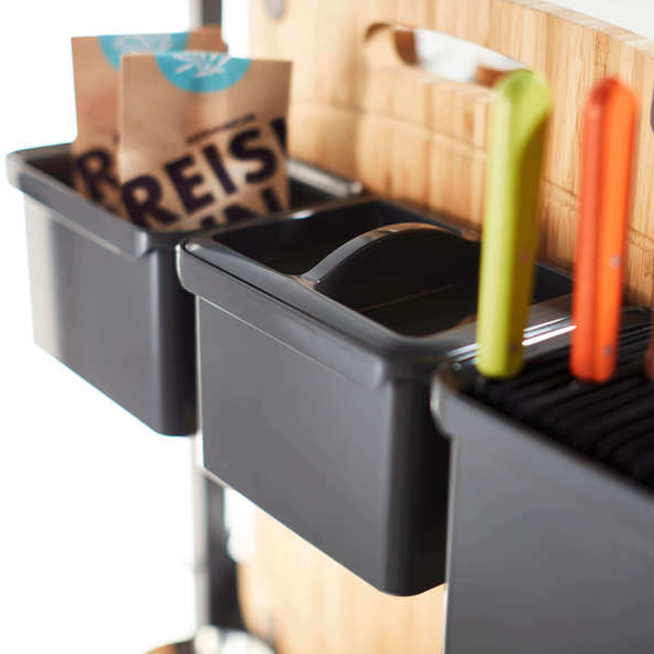 Nahaufnahme von zwei schwarzen YouboXx Einhängeboxen Set 5 für DISPENSA Apothekerschrank, die an einem Küchenwagen hängen; eine hält eine Papiertüte, die andere enthält einen Utensilien-Organizer mit Messern mit grünen und orangen Griffen. Im Hintergrund ist ein hölzernes Schneidebrett zu sehen.