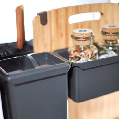 Nahaufnahme des YouboXx Einhängeboxen Set 5 für DISPENSA Apothekerschrank, mit schwarzen Behältern, einem hölzernen Schneidebrett, zwei gläsernen Gewürzdosen mit Korkdeckeln und Utensilien im Hintergrund als Teil des YouboXx-Systems.