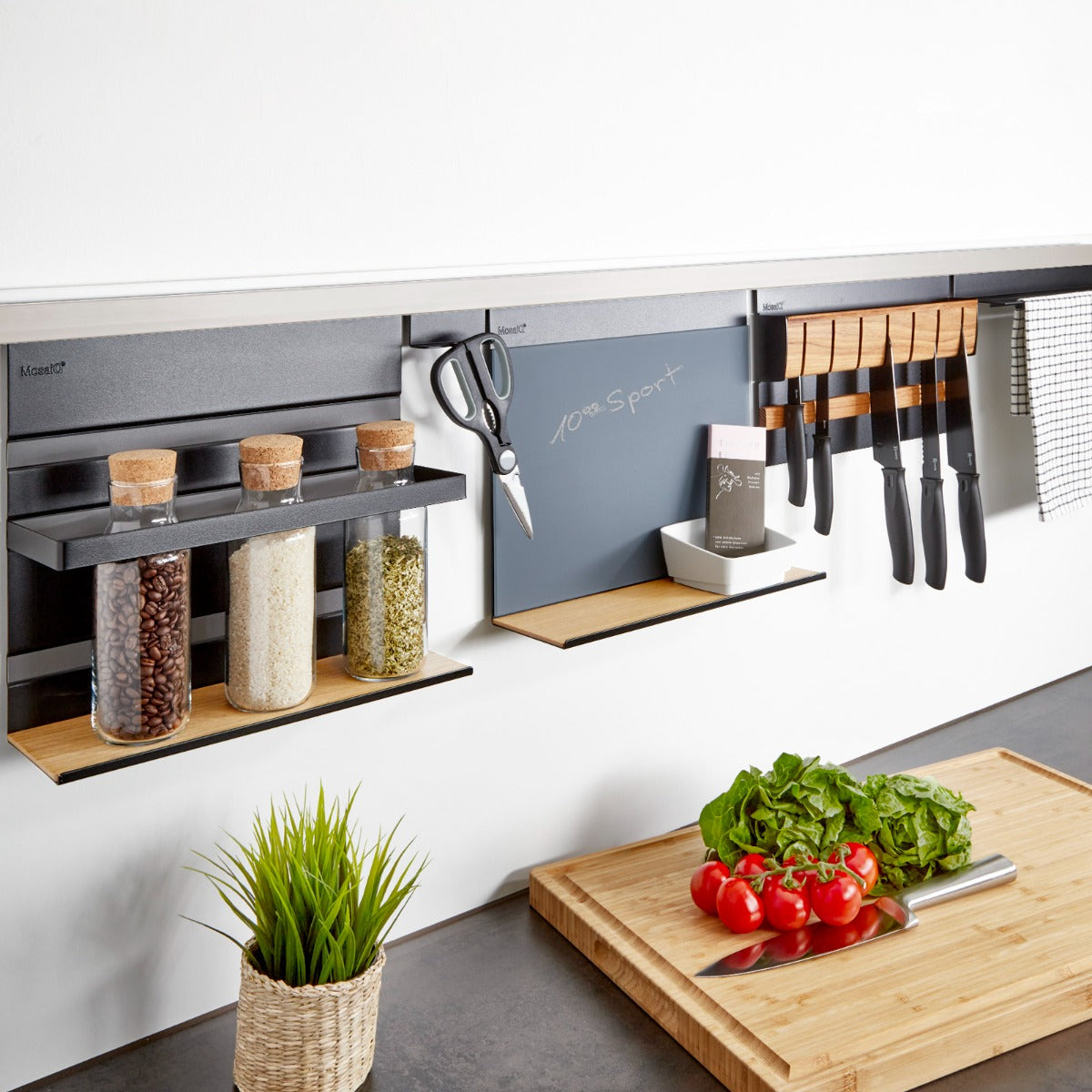 Eine moderne Küche mit Wandregalen und dem Linero MosaiQ EasyLine schwarz, Aluprofil Edelstahloptik von Kesseböhmer mit Gläsern, Scheren, Messern und einem Handtuch. Auf dem Tresen: ein Schneidebrett mit Gemüse, ein Messer und eine Topfpflanze. Die Szene ist hell und aufgeräumt.