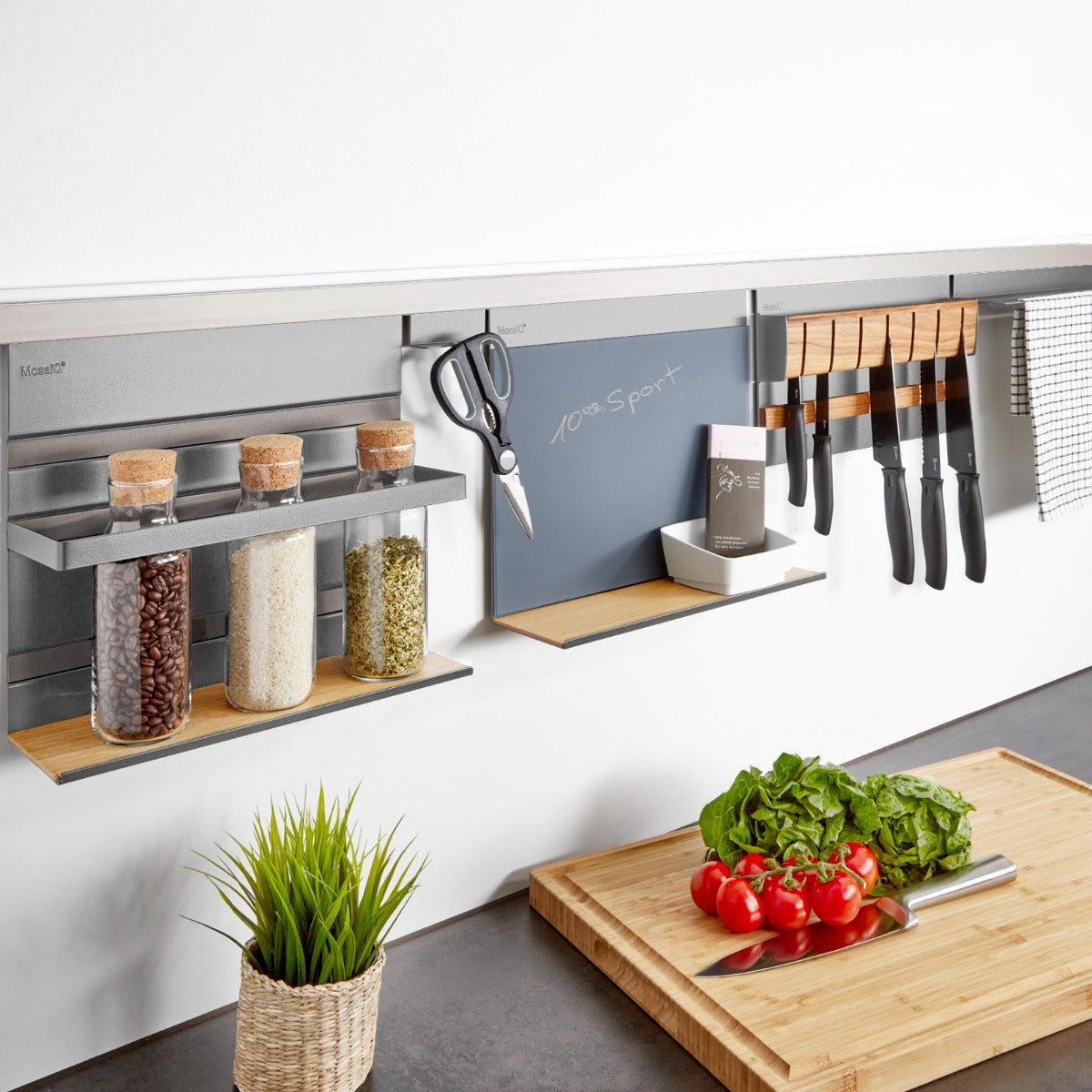 Die Linero MosaiQ EasyLine titangrau Aluprofil Edelstahloptik 600-1800 mm von Kesseböhmer ordnet Gläser, Utensilien und Messer stilvoll an der Küchenwand über einer Theke mit frischem Gemüse, Handtuch und Kreidetafel.