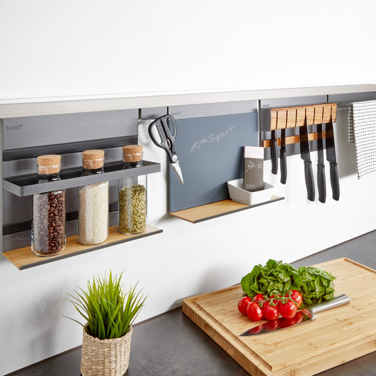 Eine moderne Küchenarbeitsplatte mit dem Linero MosaiQ EasyLine graphitschwarz, Aluprofil Edelstahloptik 600 - 1800 mm von Kesseböhmer, mit Ablagen und Halterungen für Gläser, Utensilien und Handtücher. Davor steht ein Schneidebrett mit Gemüse neben einer Topfpflanze.