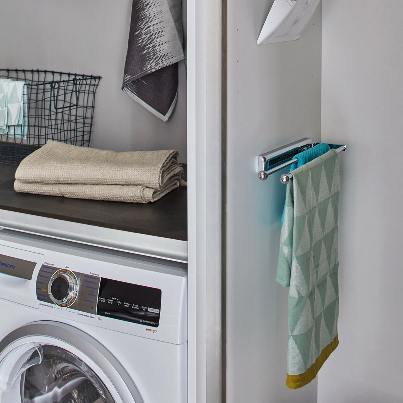 Eine moderne Waschküche mit einer platzsparenden Frontlader-Waschmaschine, ordentlich gefalteten Handtüchern auf einer dunklen Arbeitsplatte, einem Drahtkorb und einem grünen Handtuch auf dem Handtuchauszug - Handtuchhalter ausziehbar. Ideal für Küche und Bad Organisation.