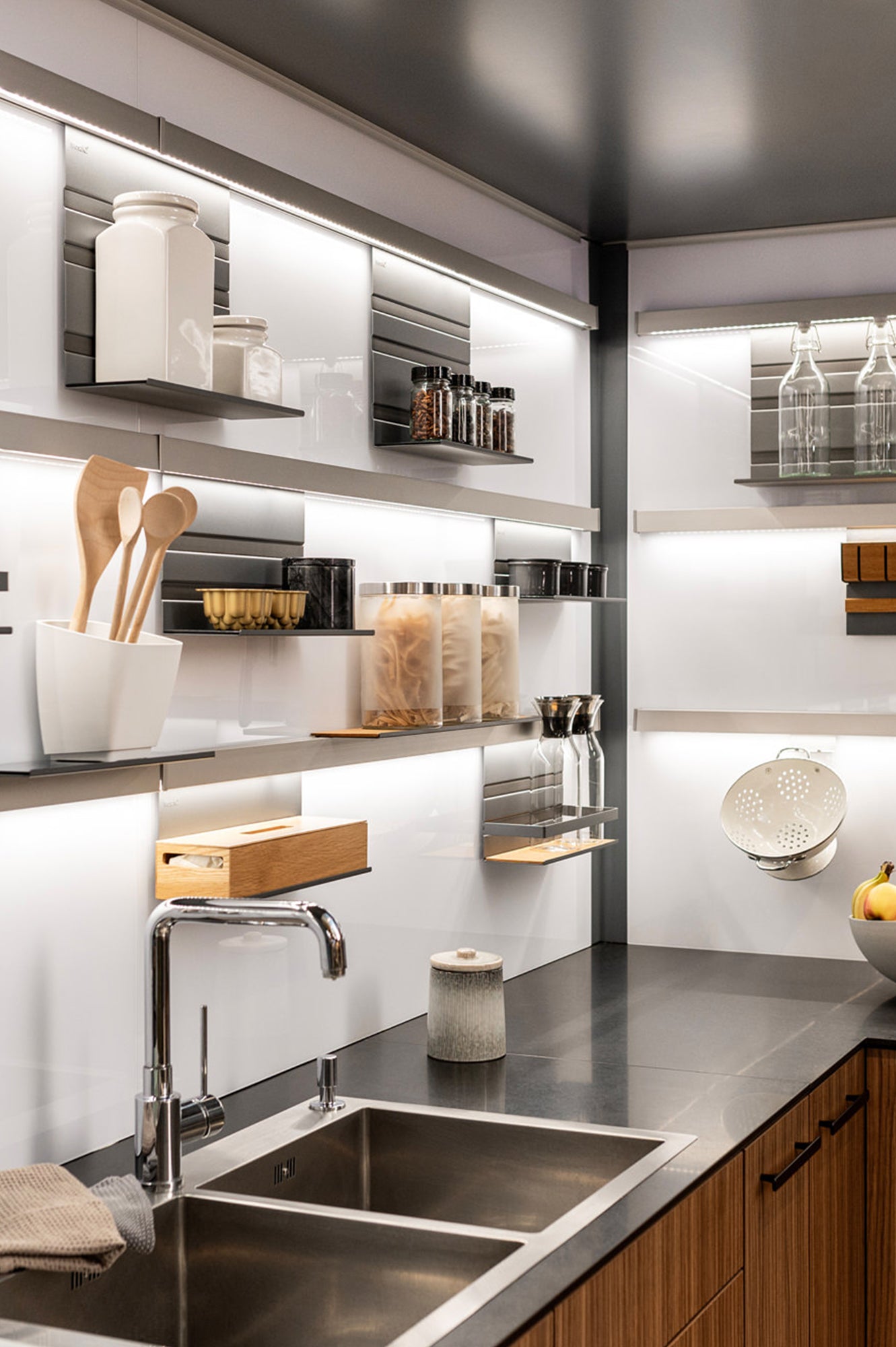 Eine moderne Küche mit Doppelspüle, schwarzer Arbeitsplatte, Holzschränken und offenen Regalen mit Gläsern, Utensilien, Gewürzen, Glasflaschen und einem Sieb bei heller, gleichmäßiger Beleuchtung.