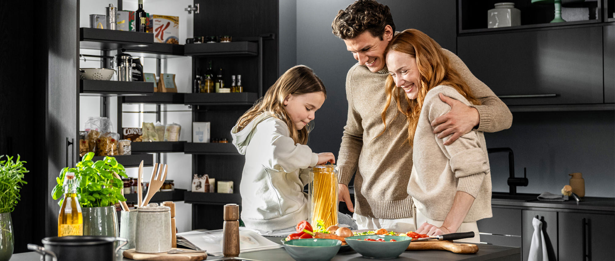 Familie (Mann, Frau, Kind) in der Küche: Kind sitzt auf der Arbeitsfläche und greift nach Spaghetti, während die Eltern sich umarmen und zusehen. Offener Vorratsschrank im Hintergrund.