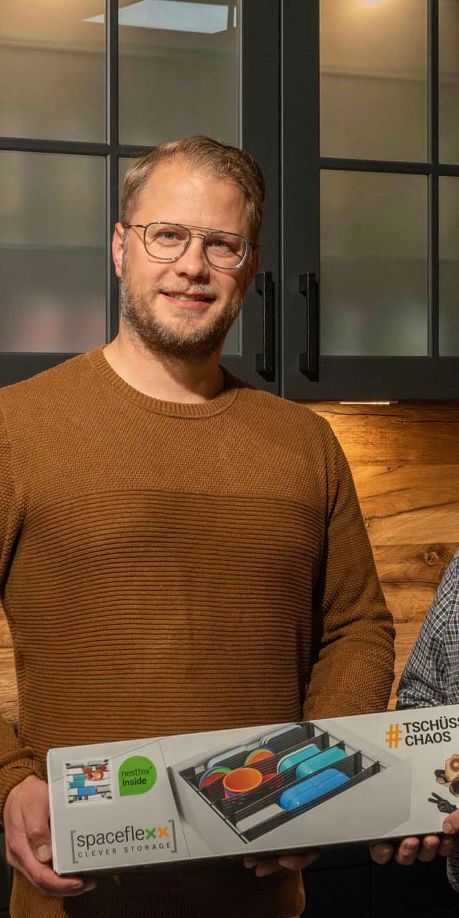 Männliche Person mit Brille steht in einer Küche und hält eine Verpackung in en Händen