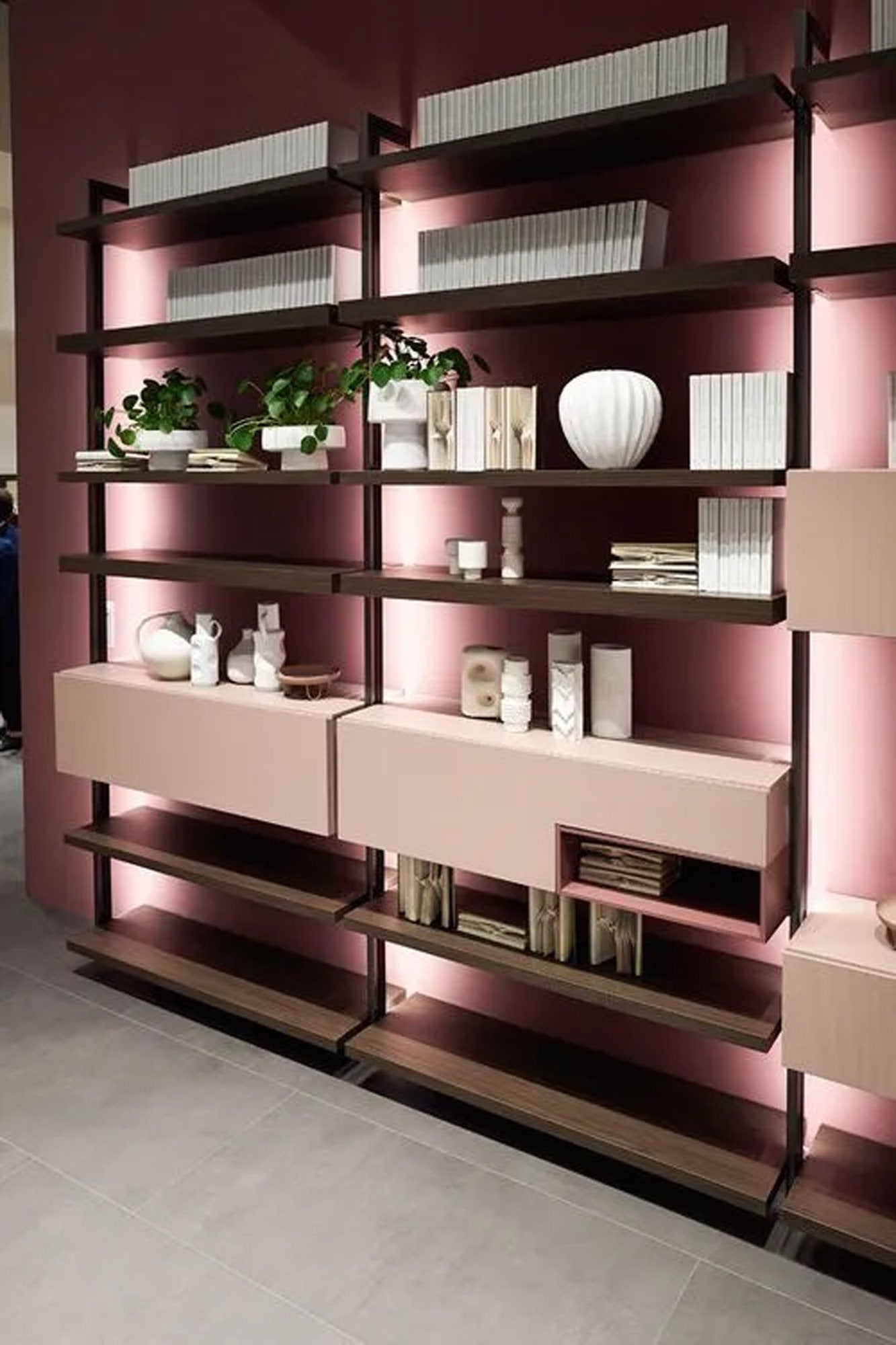 Ein modernes Regal mit dunklen Rahmen und rosafarbenen Fächern, in dem Bücher, Vasen und Dekorationsgegenstände ausgestellt sind, steht vor einer malvenfarbenen Wand mit sanfter Hintergrundbeleuchtung. Grüne Pflanzen sorgen für einen Hauch von Farbe.