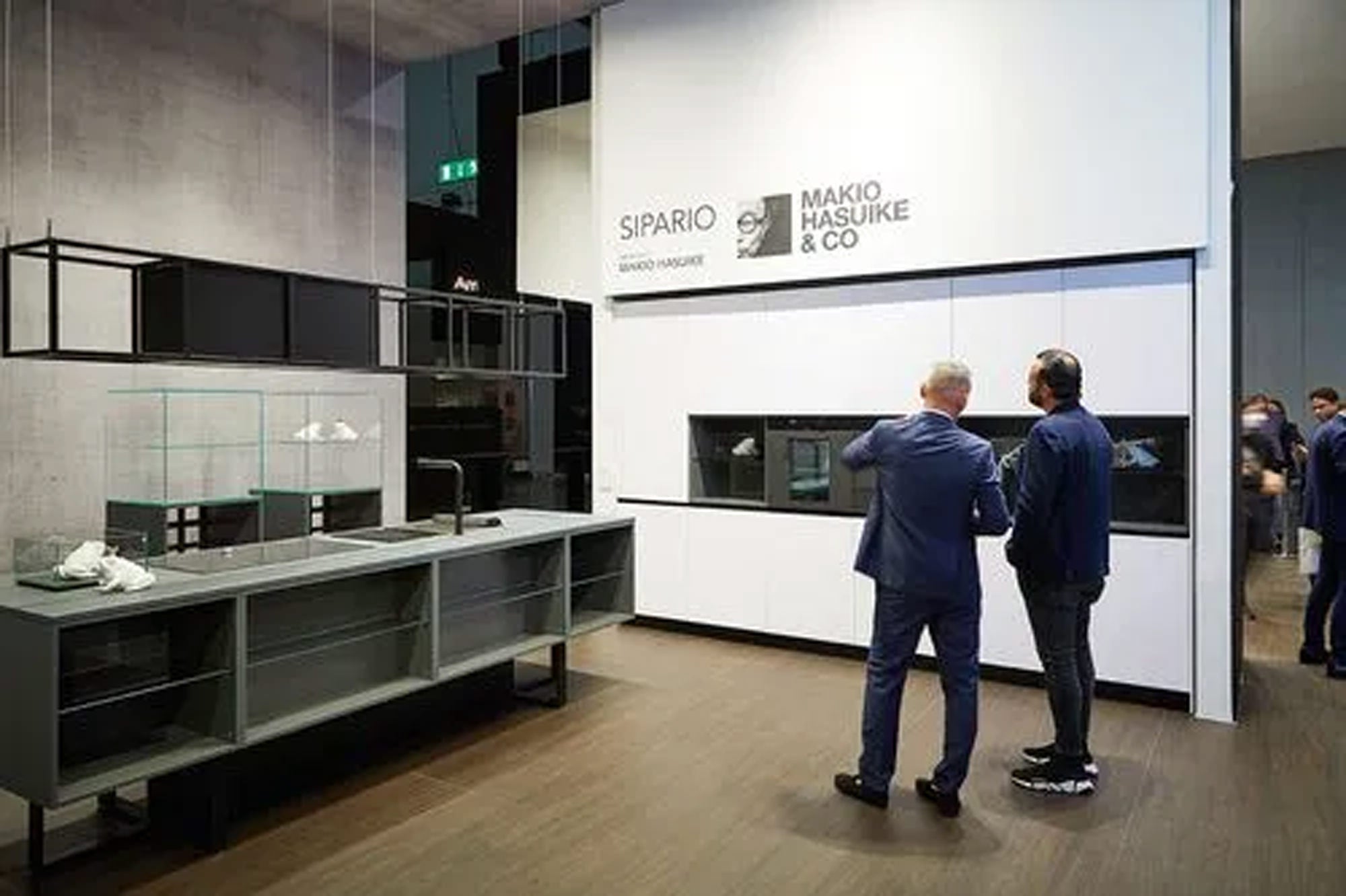 Zwei Männer in Anzügen stehen und unterhalten sich in einem modernen Ausstellungsraum mit minimalistischen Küchenauslagen. An der Wand prangt das Branding von SIPARIO und MAKIO HASUIKE & CO. Im Hintergrund sind mehrere Personen zu sehen.