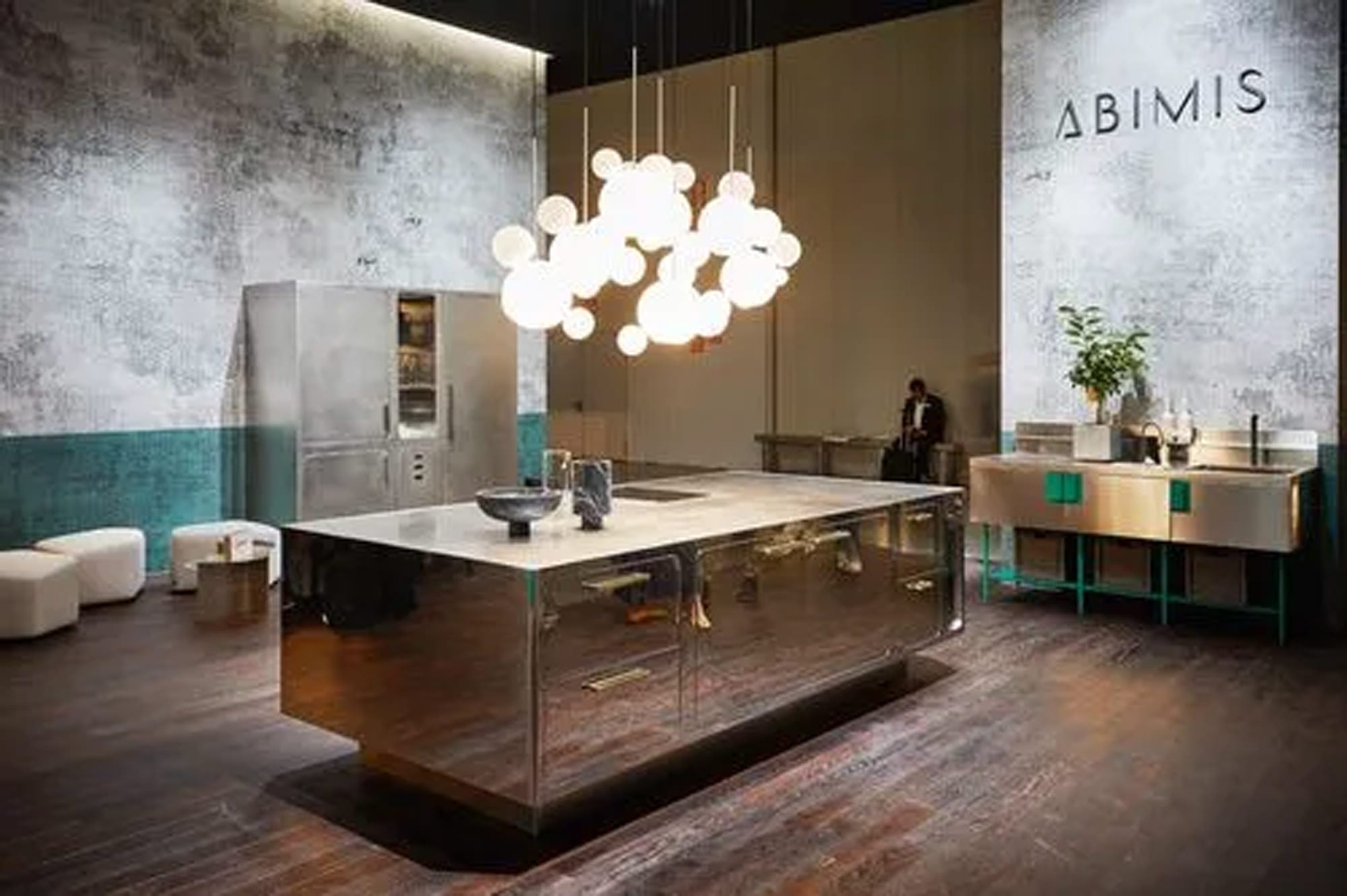 Eine moderne Küche mit einer großen Edelstahlinsel, zeitgenössischen Beleuchtungskörpern, Metallschränken, einem hohen Kühlschrank und wenigen Sitzgelegenheiten. Das Wort ABIMIS steht an der strukturierten Wand.