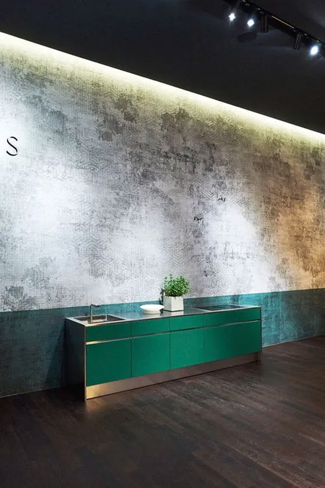 Moderne Küche mit grünen Schränken, einem Waschbecken und einer kleinen Topfpflanze vor einer strukturierten grauen Wand mit dramatischer Beleuchtung von oben. Dunkle Holzböden kontrastieren mit dem minimalistischen Design.