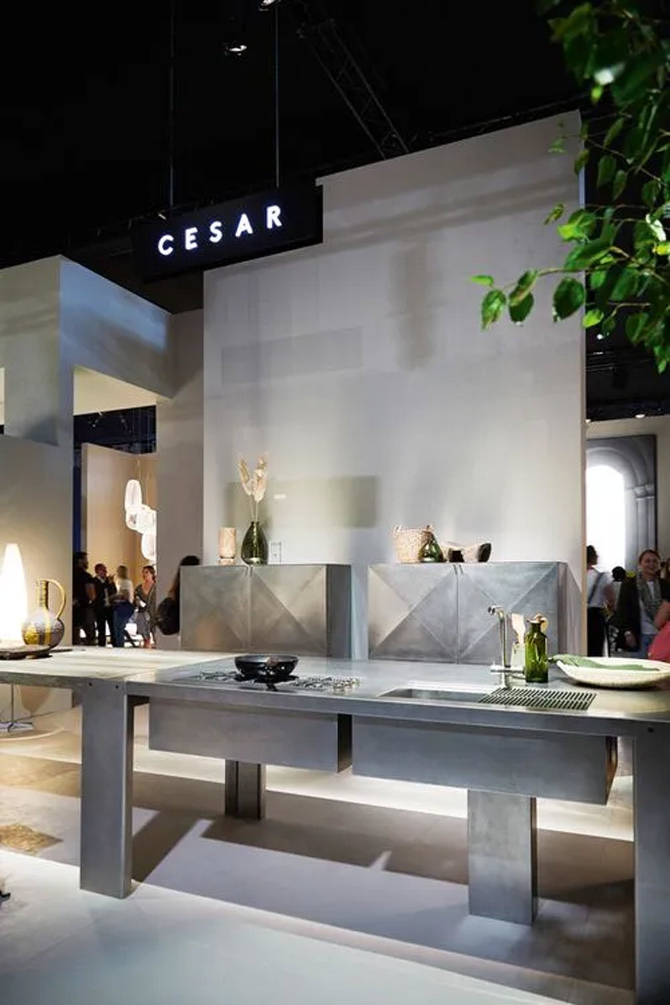 Moderner Messestand mit eleganten, minimalistischen Metallmöbeln und Dekoration unter einem Schild mit der Aufschrift CESAR. Dekorative Pflanzen und Vasen sind ausgestellt, im Hintergrund sind Menschen zu sehen.