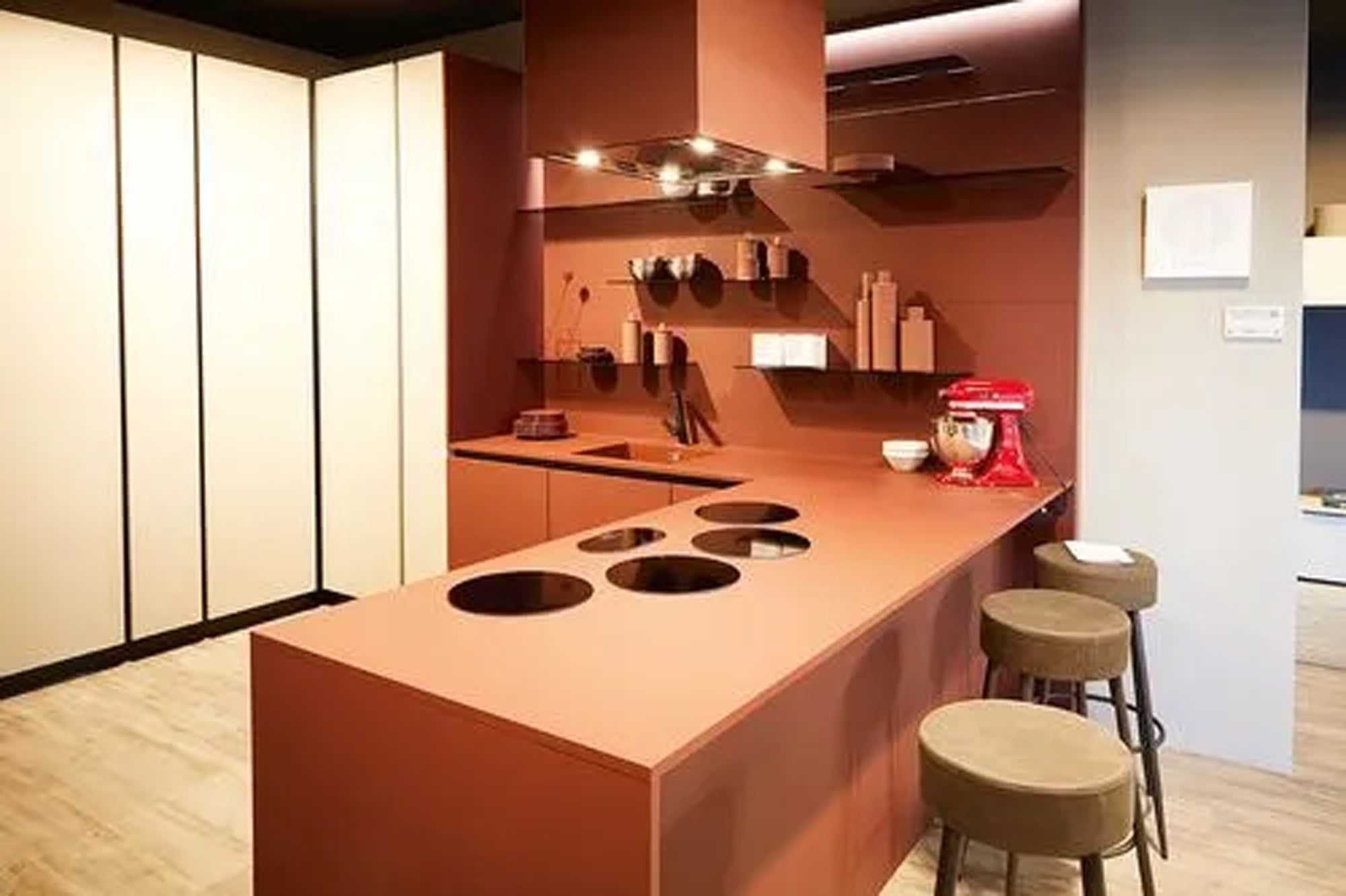 Eine moderne Küche mit mattbraunen Schränken und Arbeitsflächen, einem eingebauten Herd, drei braunen Hockern, offenen Regalen mit Geschirr und einem roten Standmixer auf dem Tresen. Der Raum hat ein minimalistisches, zeitgenössisches Design.