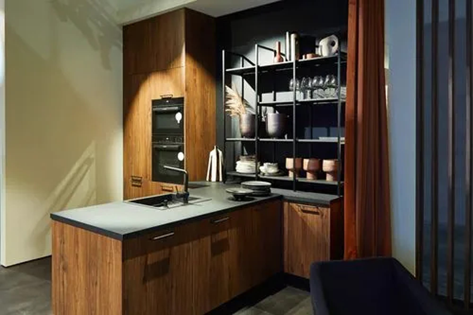 Moderne Küche mit dunklen Holzschränken, schwarzen Arbeitsplatten, einem eingebauten Backofen, offenen schwarzen Regalen für Geschirr und Dekoration und einer Spüle auf der Insel, alles in gedämpftem Licht.