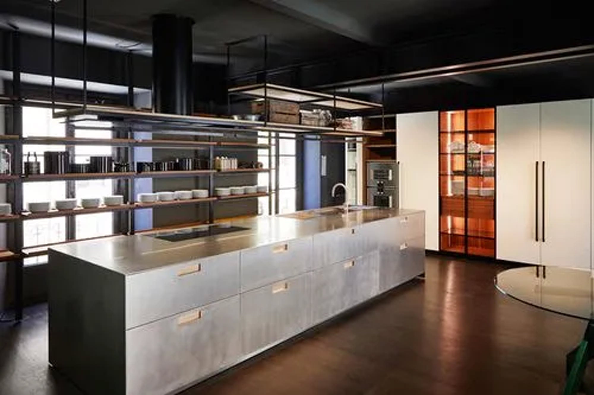 Eine moderne Küche mit einer eleganten Metallinsel, offenen Regalen mit Geschirr und Gläsern, einem eingebauten Ofen und einem großen Kühlschrank. Die warme Beleuchtung unterstreicht das minimalistische und moderne Design.