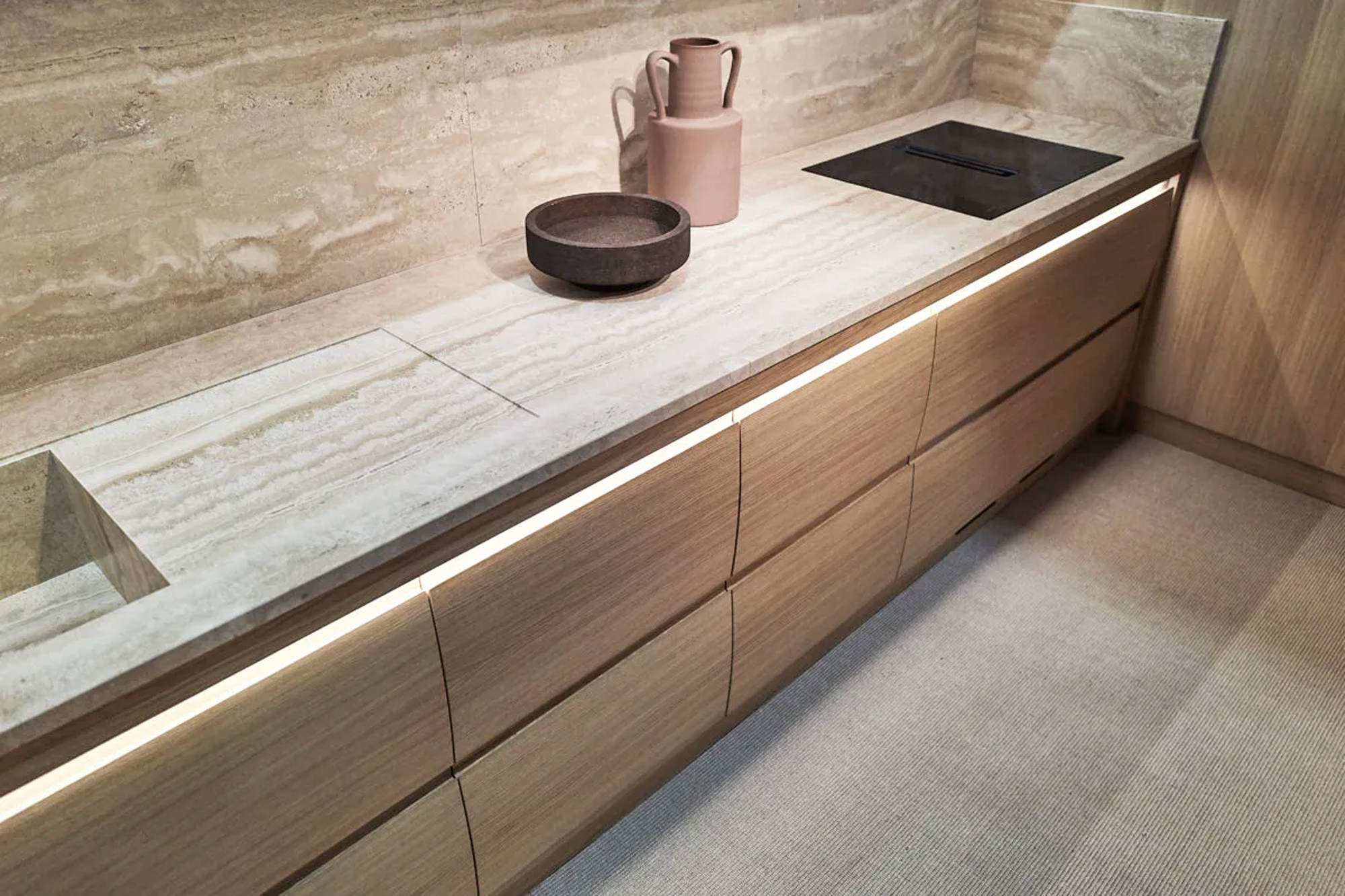 Minimalistische Küche mit hellen Holzschränken, einer beigen Arbeitsplatte aus Stein, einer integrierten Spüle, einem schwarzen Elektrokochfeld und dekorativen Töpfen, alles beleuchtet von einer Unterbauleuchte.