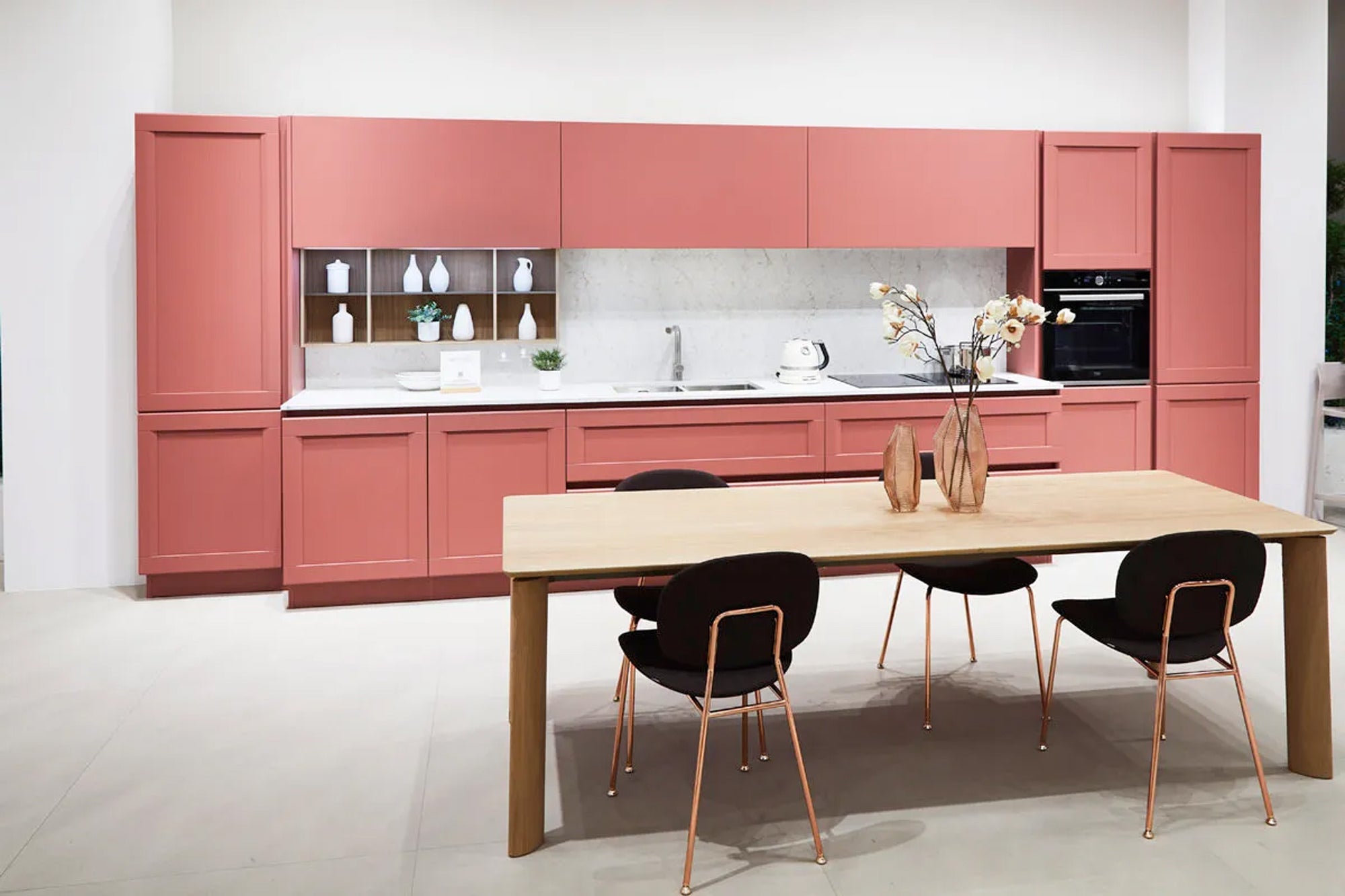 Moderne Küche mit mattrosa Schränken, weißen Arbeitsplatten, Einbaugeräten und einem Esstisch aus hellem Holz. Vier schwarze Stühle mit goldenen Beinen umgeben den Tisch, der mit zwei Vasen und Trockenblumen dekoriert ist.