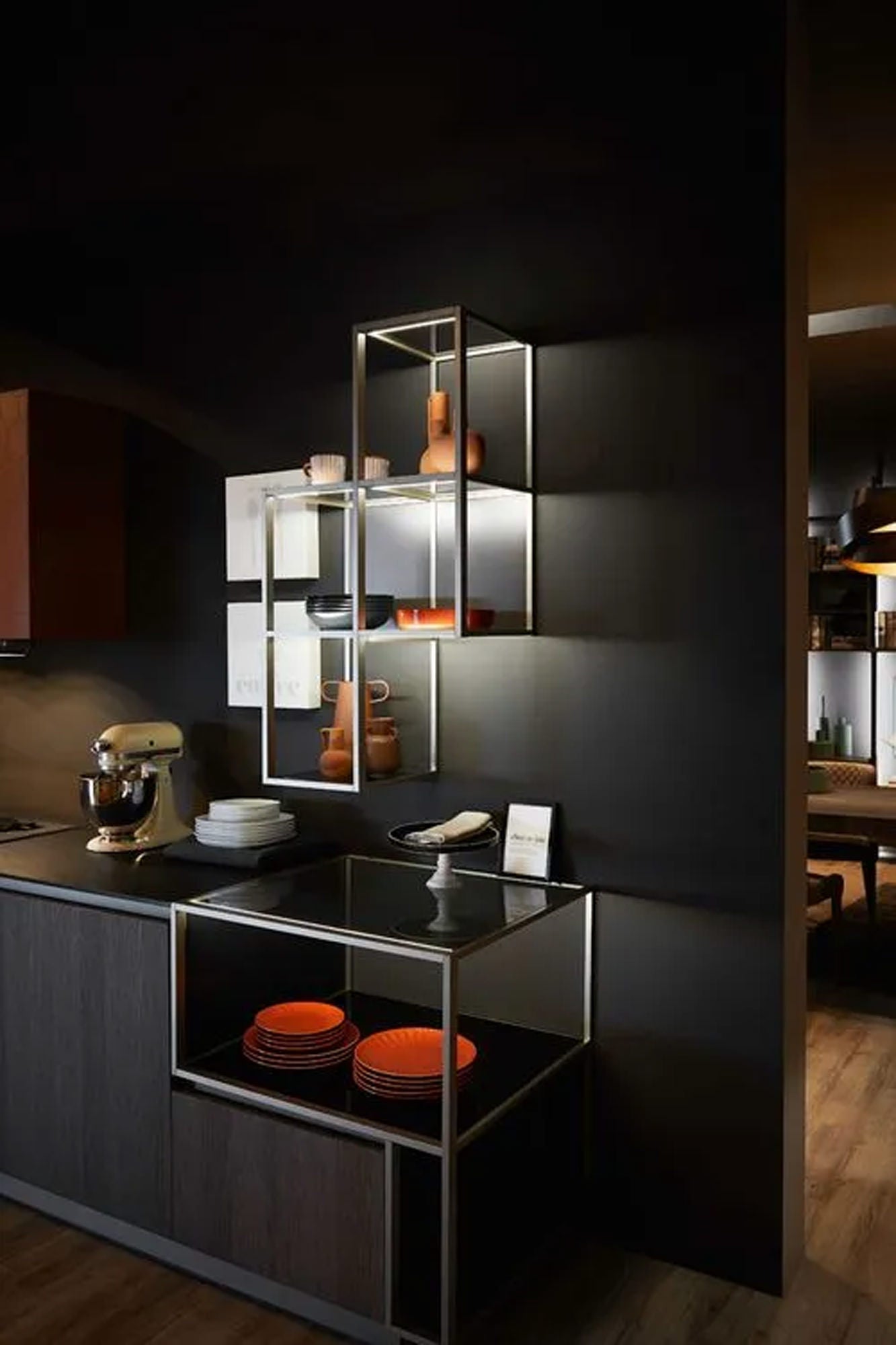 Eine moderne Küche mit dunklen Schränken, offenen Metallregalen mit orangefarbenem und neutralem Geschirr, einem Standmixer auf dem Tresen und warmer Beleuchtung schafft eine gemütliche, moderne Atmosphäre.