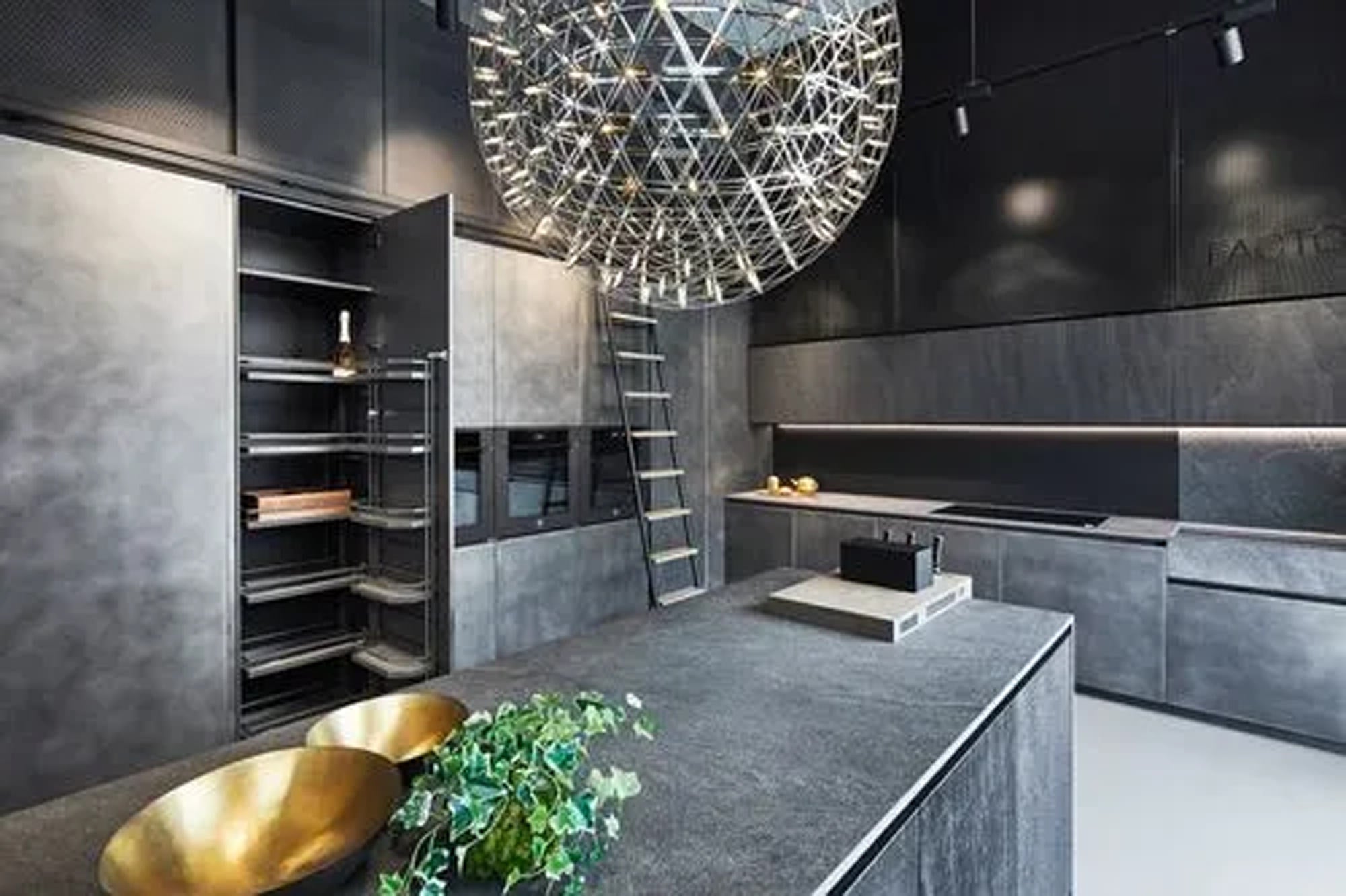 Moderne Küche mit dunkelgrauen Schränken, einer großen Insel, offenen Regalen, einem kugelförmigen Metallkronleuchter, einer hohen Leiter, goldenen Schalen und einer kleinen grünen Pflanze auf der Insel. Der Raum hat ein schlichtes, industrielles Design.