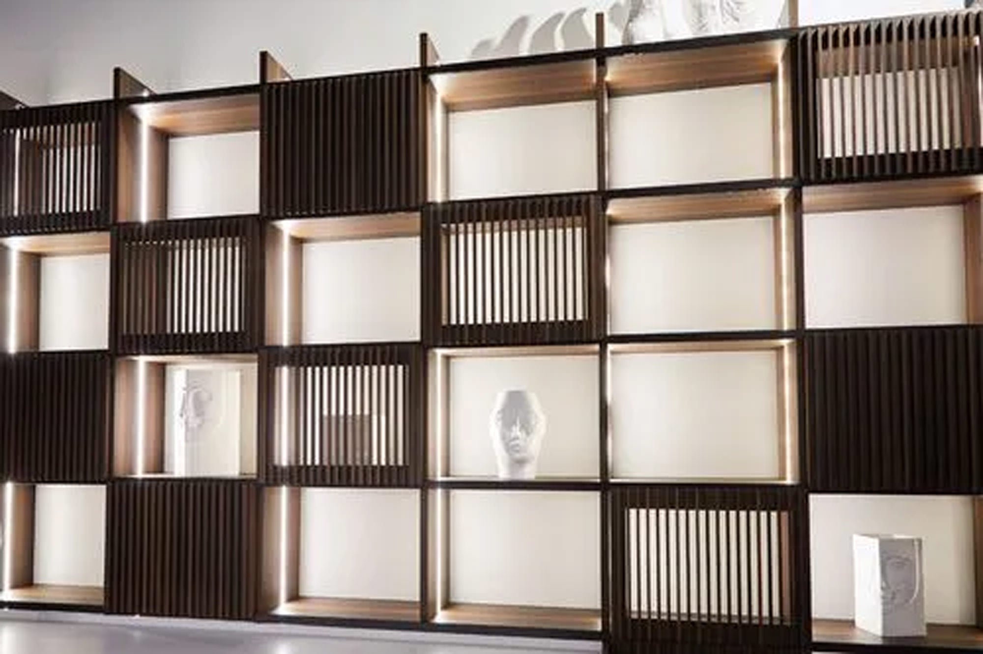 Ein modernes Wandregal, bei dem sich offene und geschlossene Bereiche abwechseln, mit Holzlatten und beleuchteten Regalböden, auf denen weiße Kopfbüsten und minimalistische Dekorationsstücke stehen.