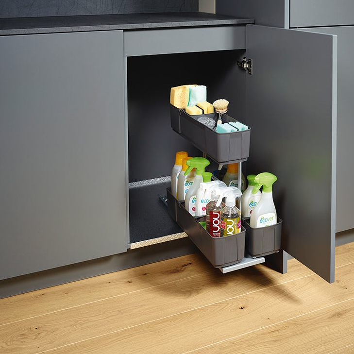 Ein grauer Küchenschrank mit geöffneter Tür zeigt den cleaningAGENT - Putzschrank Organizer & Putzmittelauszug, in dem Sprühflaschen, Schwämme und Bürsten auf zwei Regalböden über einem Holzboden stehen.