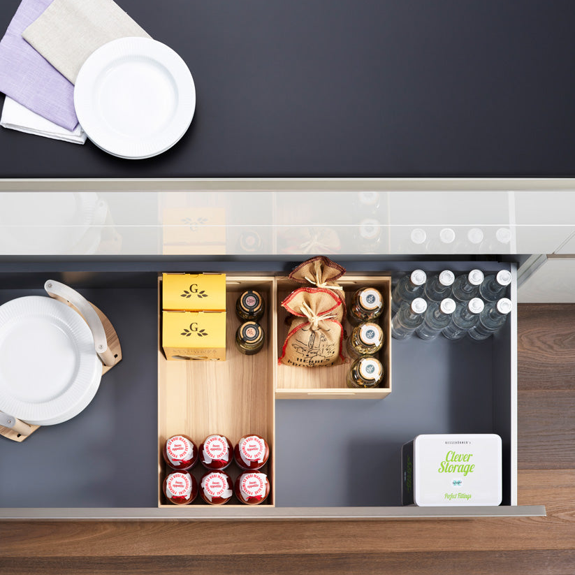 Eine Küchenschublade mit Papptellern, die FineLine Holzbox mit Grifflöchern für Flaschengetränke und verpackte Waren, ein Clever Storage Container und Wasser in Flaschen. Servietten und ein Teller auf der Theke - Design und Stauraum in Perfektion.