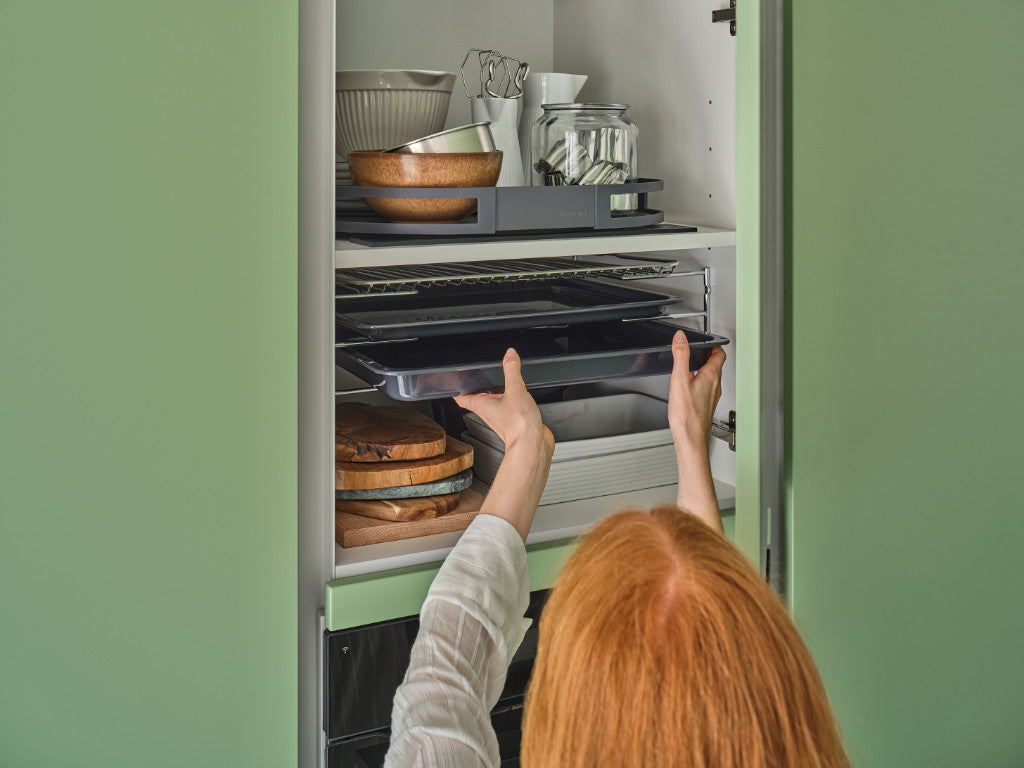 Frau, deren Kopf nur von hinten zu sehen ist, greift nach einem Backblech aus einem hellgrünen Küchenschrank. Küchenschrank mit weiteren Utensilien gefüllt.