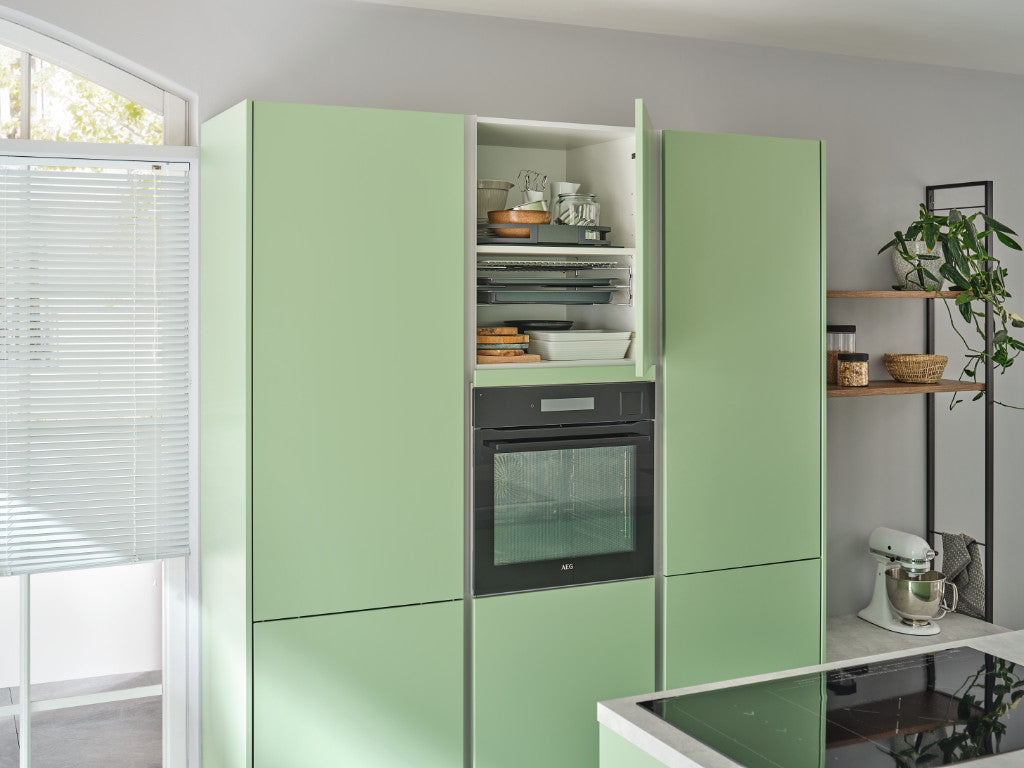Geöffneter, hellgrüner Küchenhochschrank mit Backformen, Utensilien und eingebautem Backofen. Rechts davon ein Regal mit einer Küchenmaschine. Links ein Fenster.