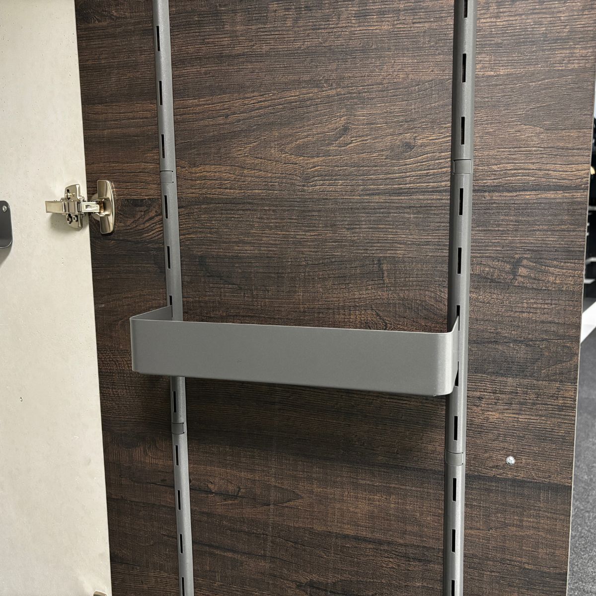 Eine Schranktür aus Holz mit einer Montageschiene aus Metall und einer seitlichen Ablage von K-Line, die vertikal angebracht ist und über verstellbare Schlitze zur individuellen Anpassung verfügt. Das Schrankscharnier ist auf der linken Seite sichtbar.