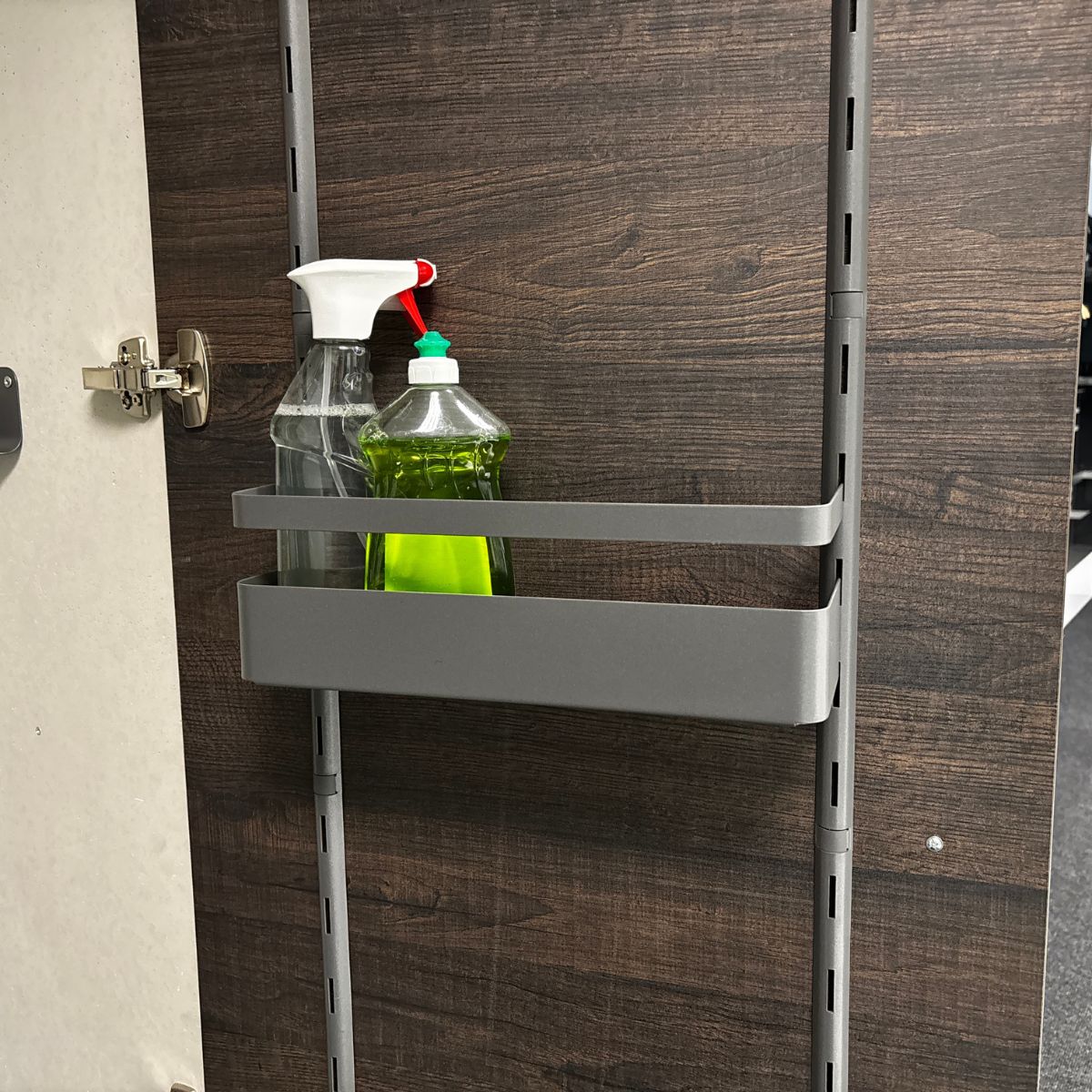 Eine K-Line Seitenablage mit Reling aus Metall, montiert an einer dunklen Holztür, hält eine Sprühflasche und eine grüne Spülmittelflasche. Das Türscharnier befindet sich links.