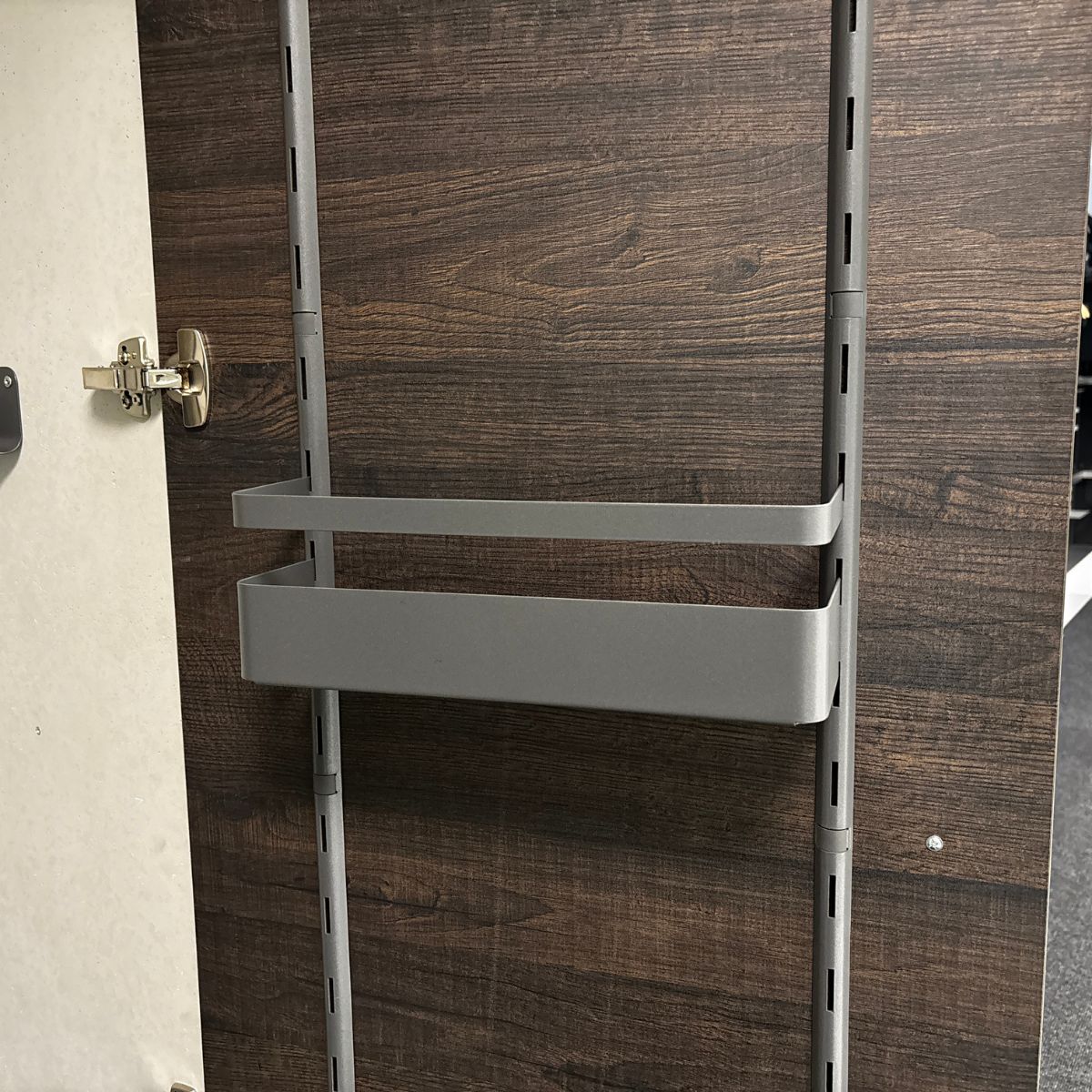 Die Schranktür aus dunklem Holz hat ein silbernes Scharnier und ein vertikales Reling-System mit der K-Line Seitenablage mit Reling, die einen rechteckigen grauen Ablagekorb hält.