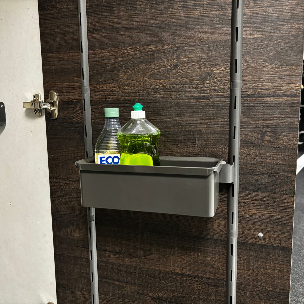 Die K-Line seitliche Trägerleiste mit Box in Grau wird an einer vertikalen Schiene befestigt, um Ihren Küchenschrank zu organisieren und Geschirrspülmittel, eine Flasche mit ECO-Label und einen grünen Schwamm vor einer dunklen, holzgemusterten Schrankwand ordentlich zu verstauen.