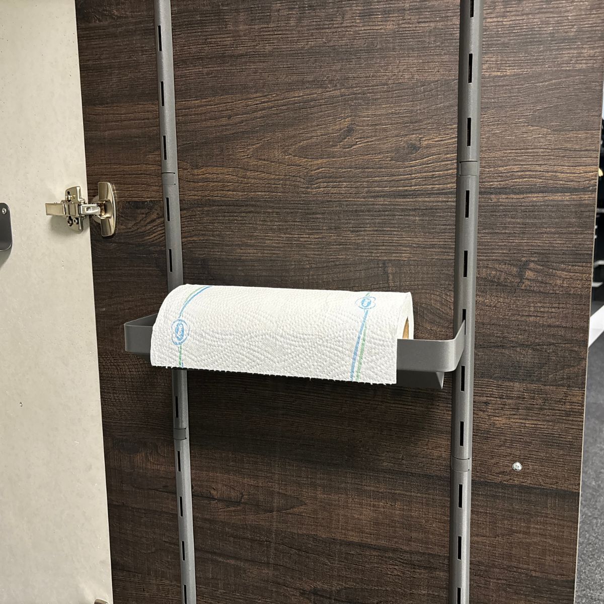 Die K-Line side Papierrollenablage hält eine Küchenrolle auf einer horizontalen Halterung, die an einer dunklen Holzplatte mit verstellbaren Regalfächern befestigt ist - ideal für die Organisation Ihrer Küche.