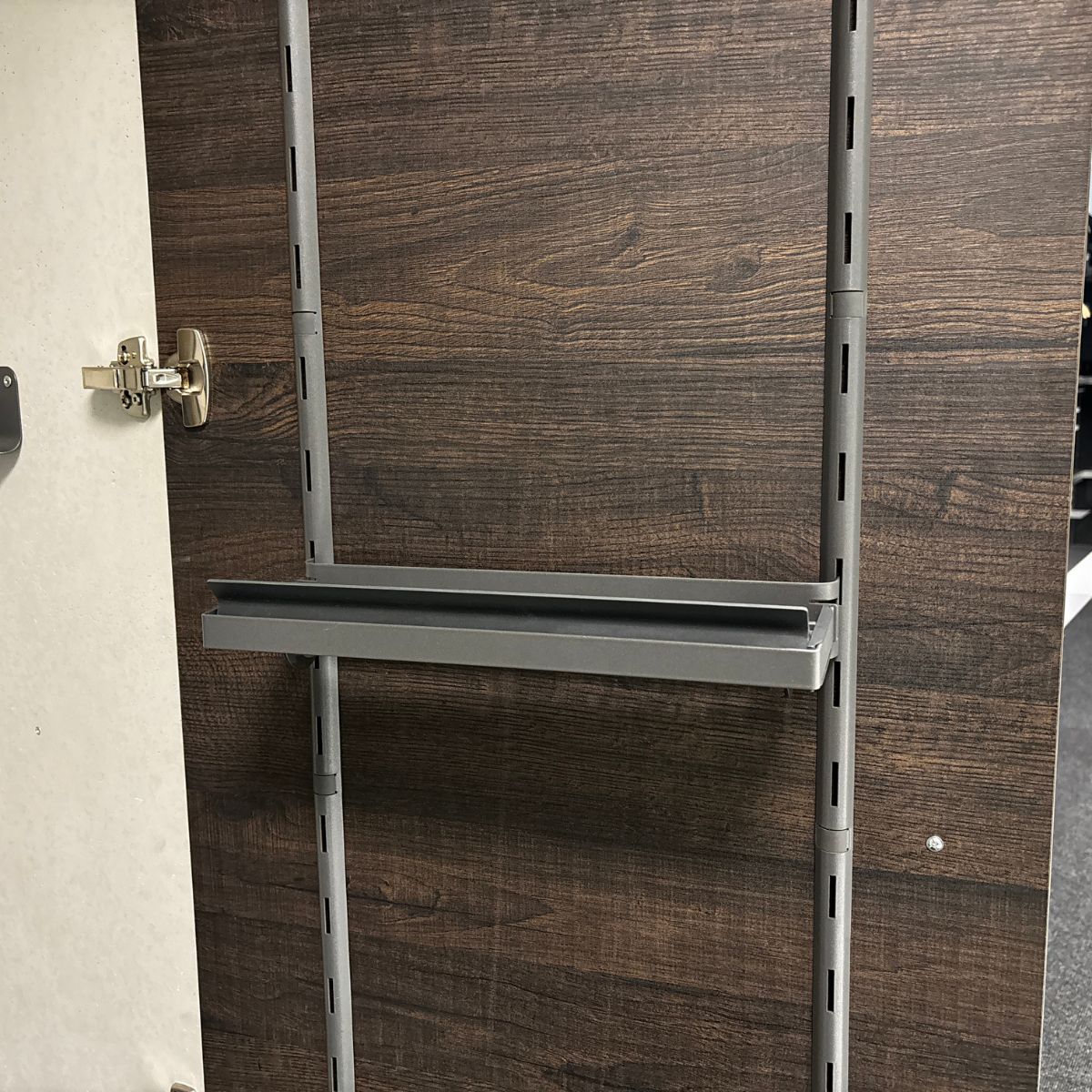 Die K-Line Side ROLLINERO - Rollenablage ist eine Metallablage, perfekt für die Folienablage, montiert auf vertikalen Schienen innerhalb einer dunklen Holzschranktür mit einem sichtbaren Metallscharnier auf der linken Seite.