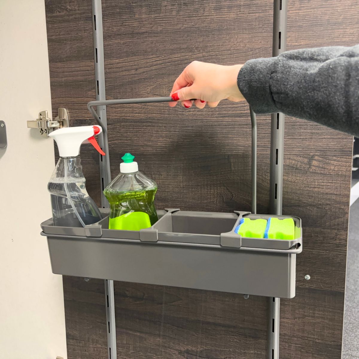Eine Hand ergreift den Griff einer grauen, an der Wand hängenden K-Line Side Tragebox, in der eine Sprühflasche, Spülmittel, zwei Schwämme und ein leeres Fach untergebracht sind - ideal, um Reinigungsmittel mühelos zu organisieren.