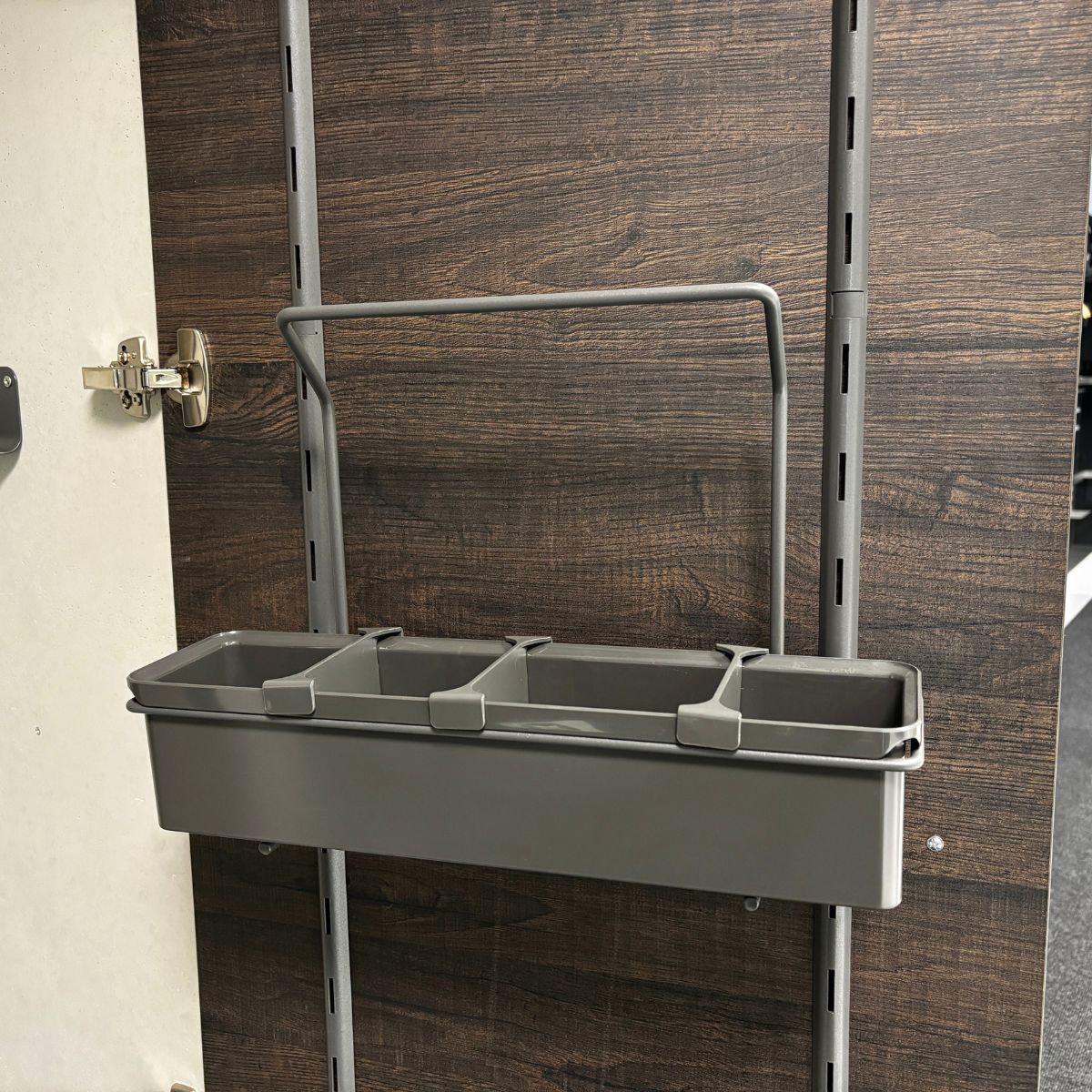 Die K-Line Side Tragebox, ein grauer, rechteckiger Hängeorganizer mit vier Fächern, wird mit einem Metallregalsystem an einer dunklen, holzgemusterten Wand montiert - ideal, um Putzmittel zu verstauen und echte Ordnung zu erleben. Auf der linken Seite befindet sich ein Schrankscharnier.