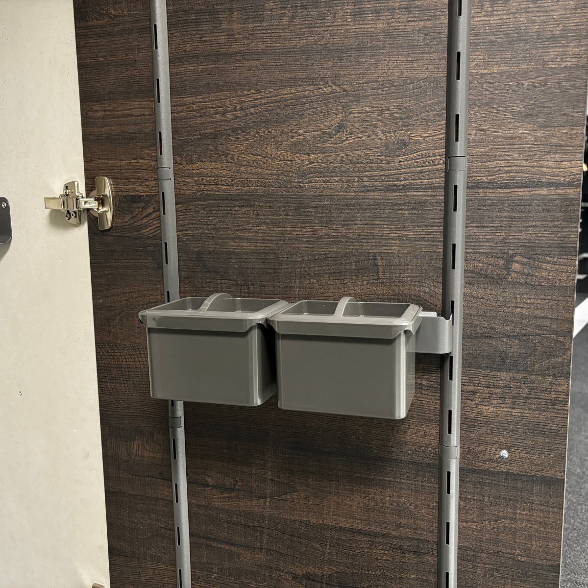 Die K-Line Seitenträgerleiste mit zwei Boxen in Grau bietet Stauraum Kleinteile und ist auf einer Metallschiene montiert, die an einer vertikalen Holzplatte mit dunkler Holzmaserung befestigt ist; links ist ein Schrankscharnier sichtbar.