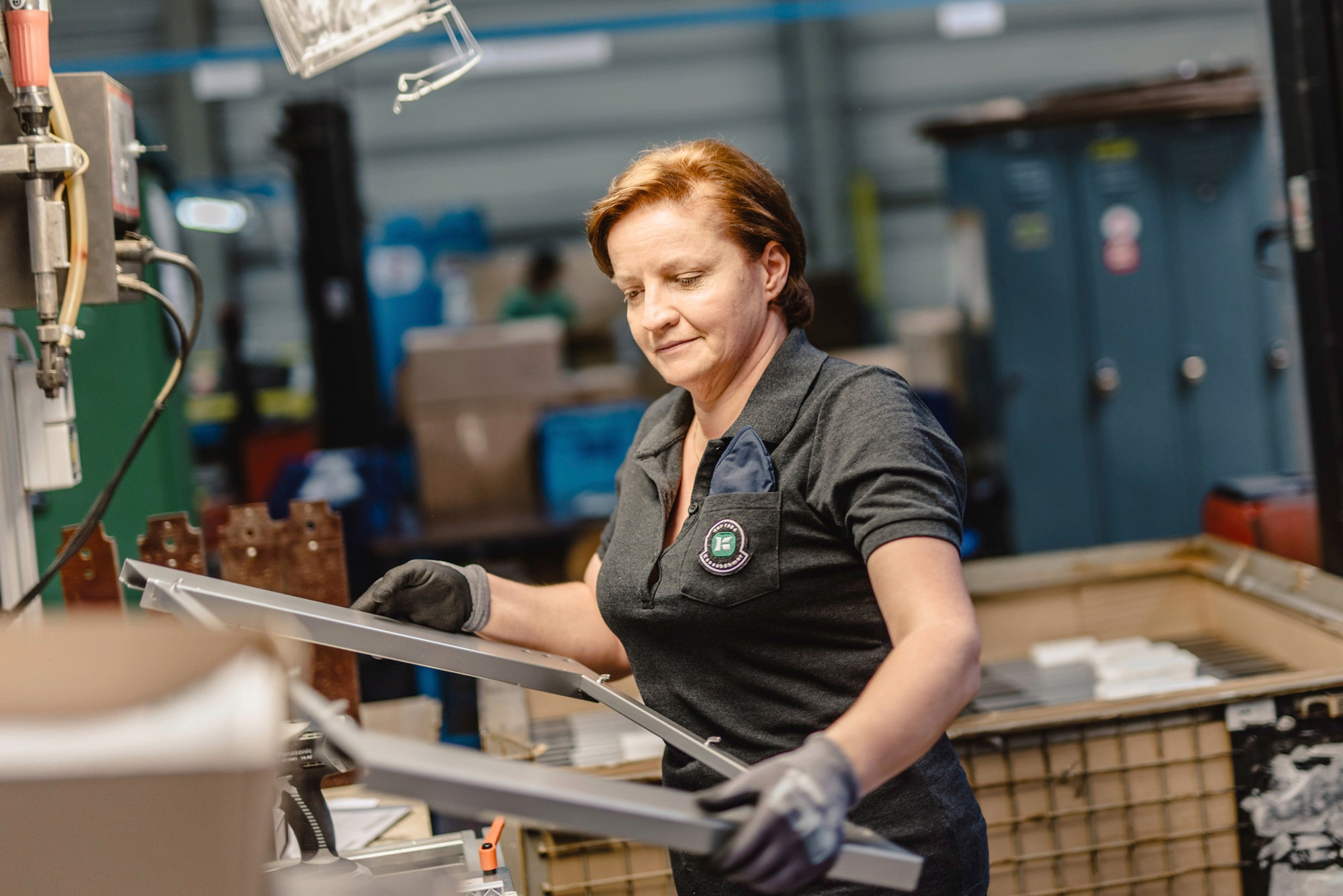Arbeiterin in Produktionshalle trägt Metallrahmen in den Händen.