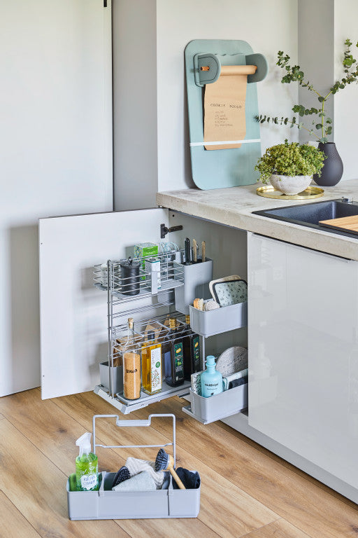 Geöffneter Unterschrank mit herausgezogenem Putzmittelauszug. Auf dem Fußboden daneben ein herausnehmbarer Korb mit bunten Reinigungsflaschen.