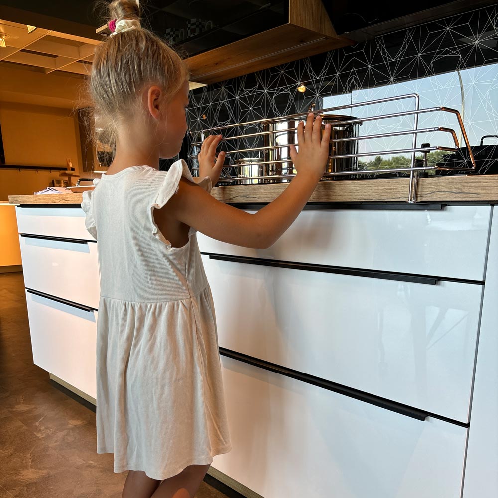 Ein junges Mädchen in einem weißen Kleid steht in einer modernen Küche und berührt ein Metallregal auf einer Arbeitsplatte mit weißen Schubladen und einer geometrischen schwarzen Aufkantung, die von der Herd & Kochfeld Kindersicherung - Herdschutzgitter geschützt wird.