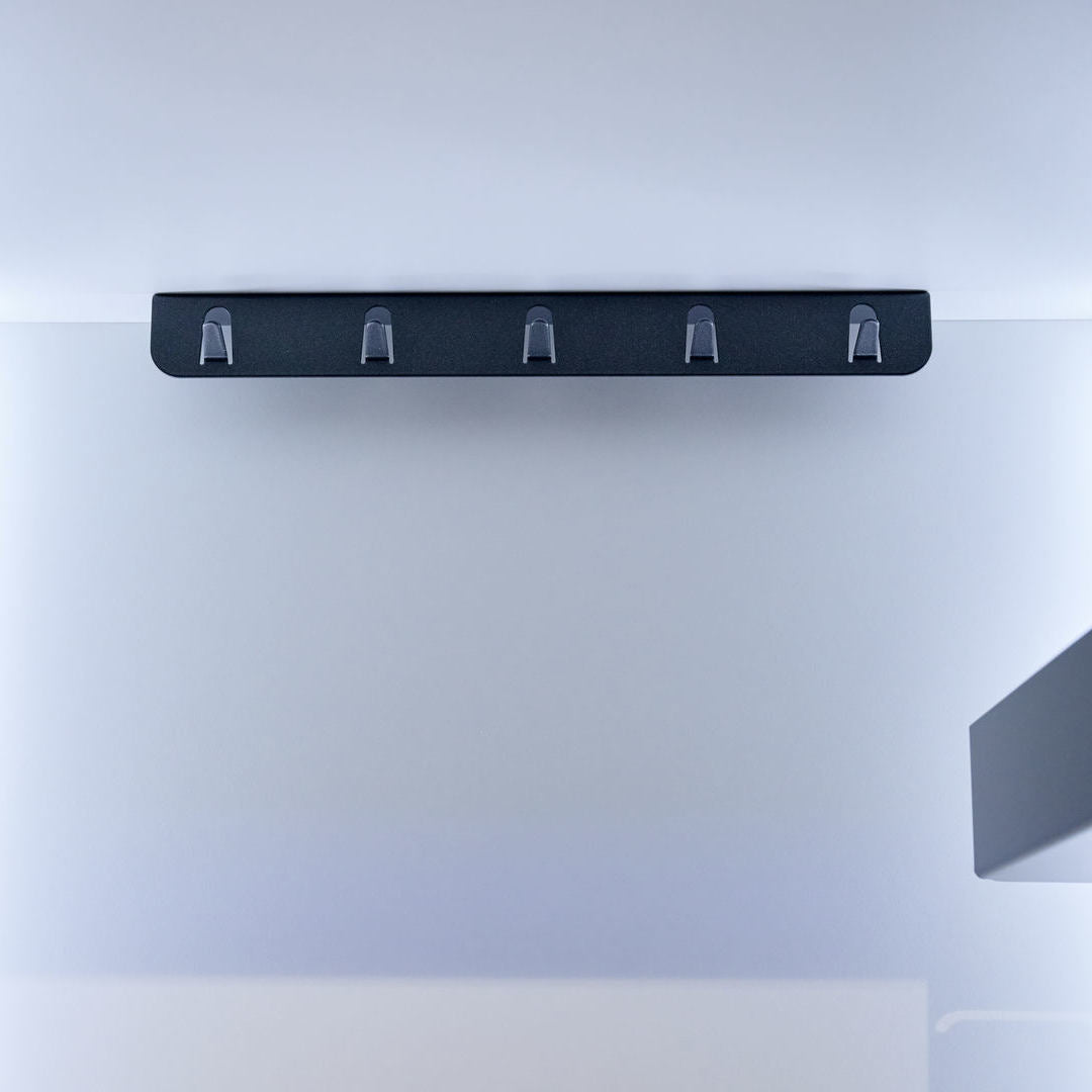 Eine minimalistische schwarze K-Line Seitenhakenleiste - Deckenbefestigung mit fünf Haken ist platzsparend aufgehängt an einer leeren weißen Wand in einem modernen, hell erleuchteten Raum.