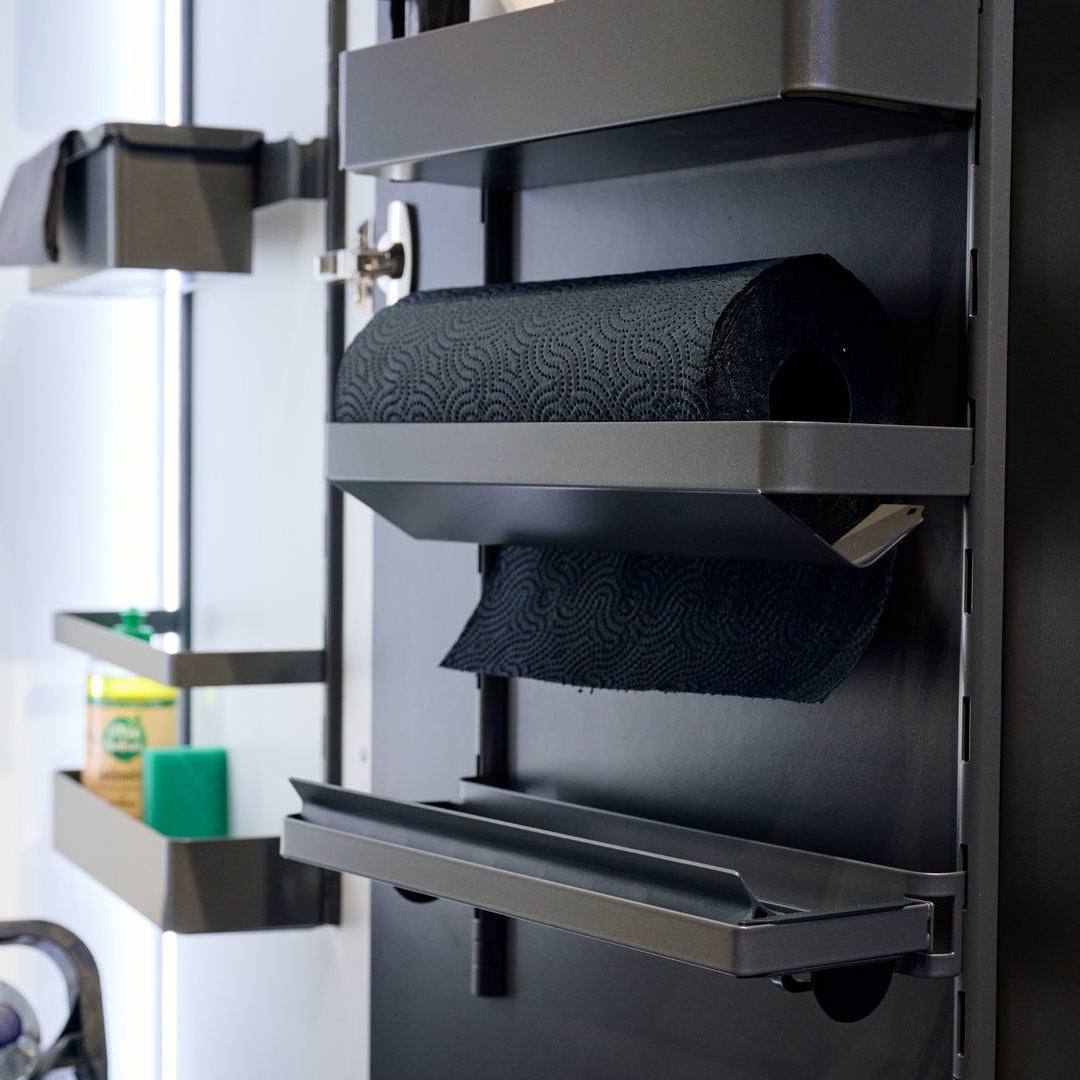 Eine Nahaufnahme der modernen K-Line Seitenpapierrollenablage mit einer schwarzen Papierhandtuchrolle. In den benachbarten Regalen befinden sich Reinigungsmittel wie ein grüner Schwamm und eine Flasche im Hintergrund.